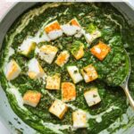 A pan filled with creamy spinach curry and cubed paneer, garnished with cream and herbs, with a gold-handled spoon resting inside.