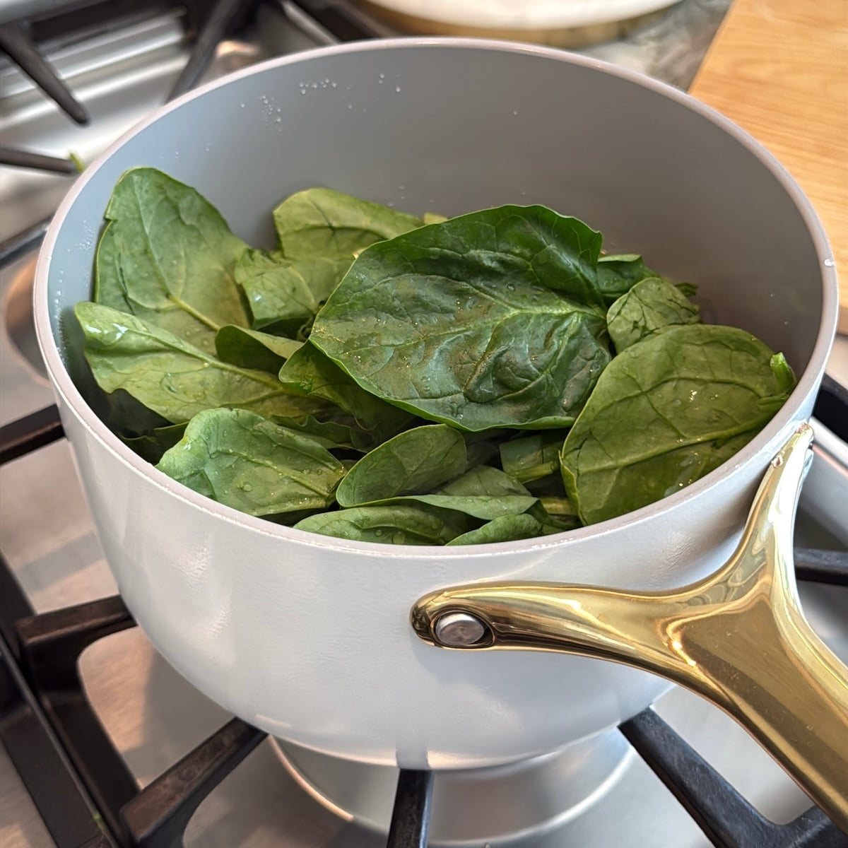 Fresh spinach leaves in a white saucepan with a gold handle on a gas stove, ready to be cooked.