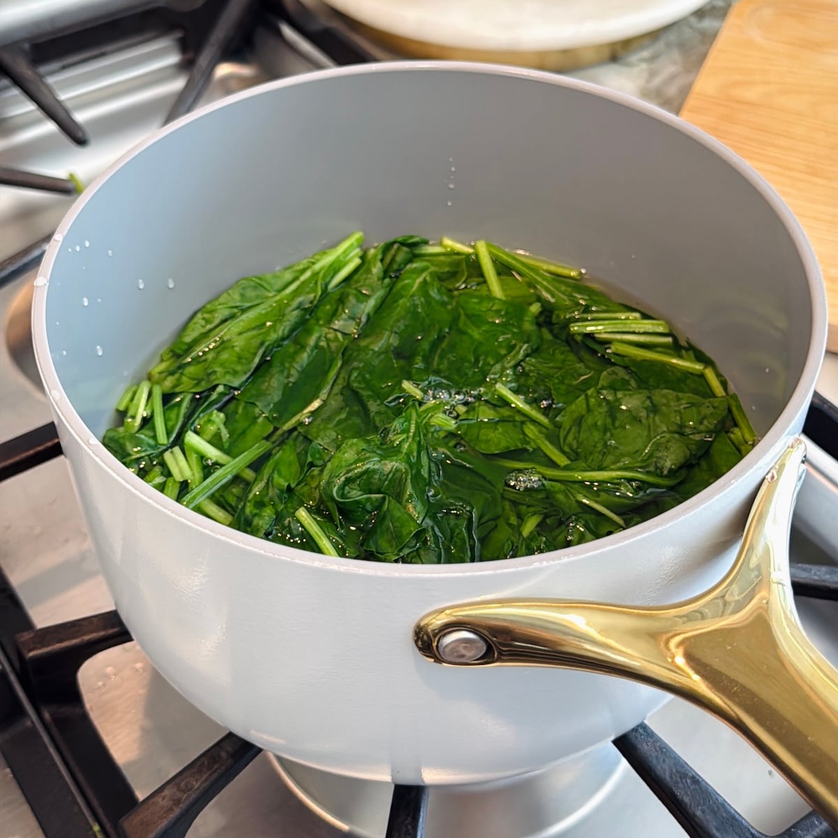 Fresh spinach leaves are being blanched in a pot of hot water on a stove, with green stems visible in the pot.