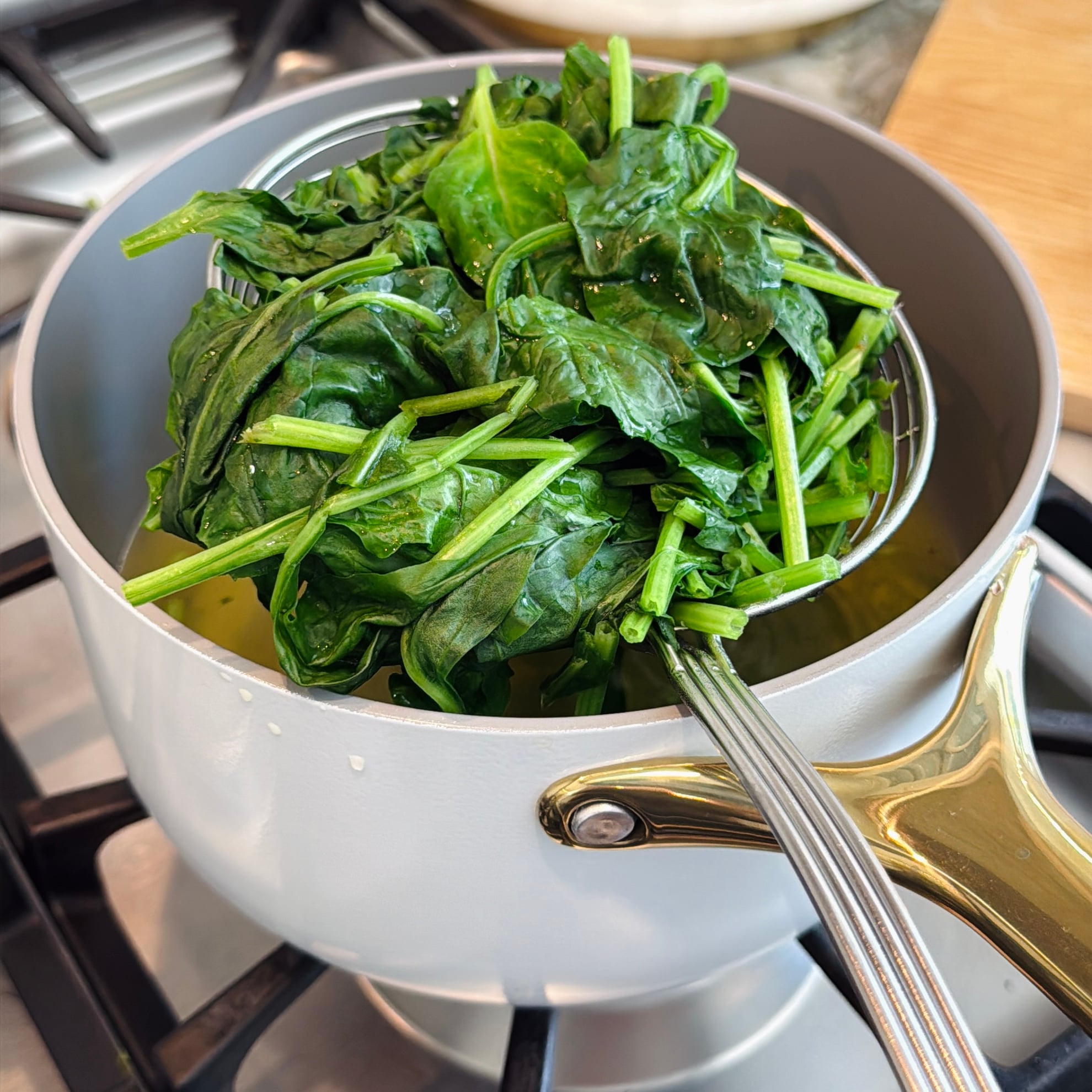 A slotted spoon holds wilted spinach above a saucepan on a stove.