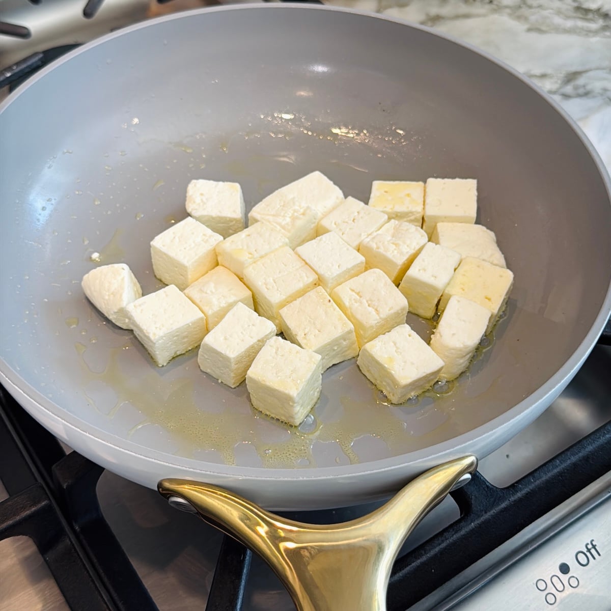 Cubes of paneer cooking in a light gray nonstick pan with oil on a stove.