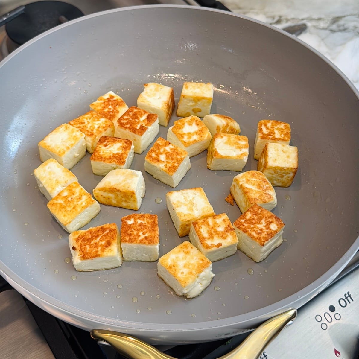 Cubed pieces of paneer are being pan-fried in a nonstick skillet, showing golden-brown edges.