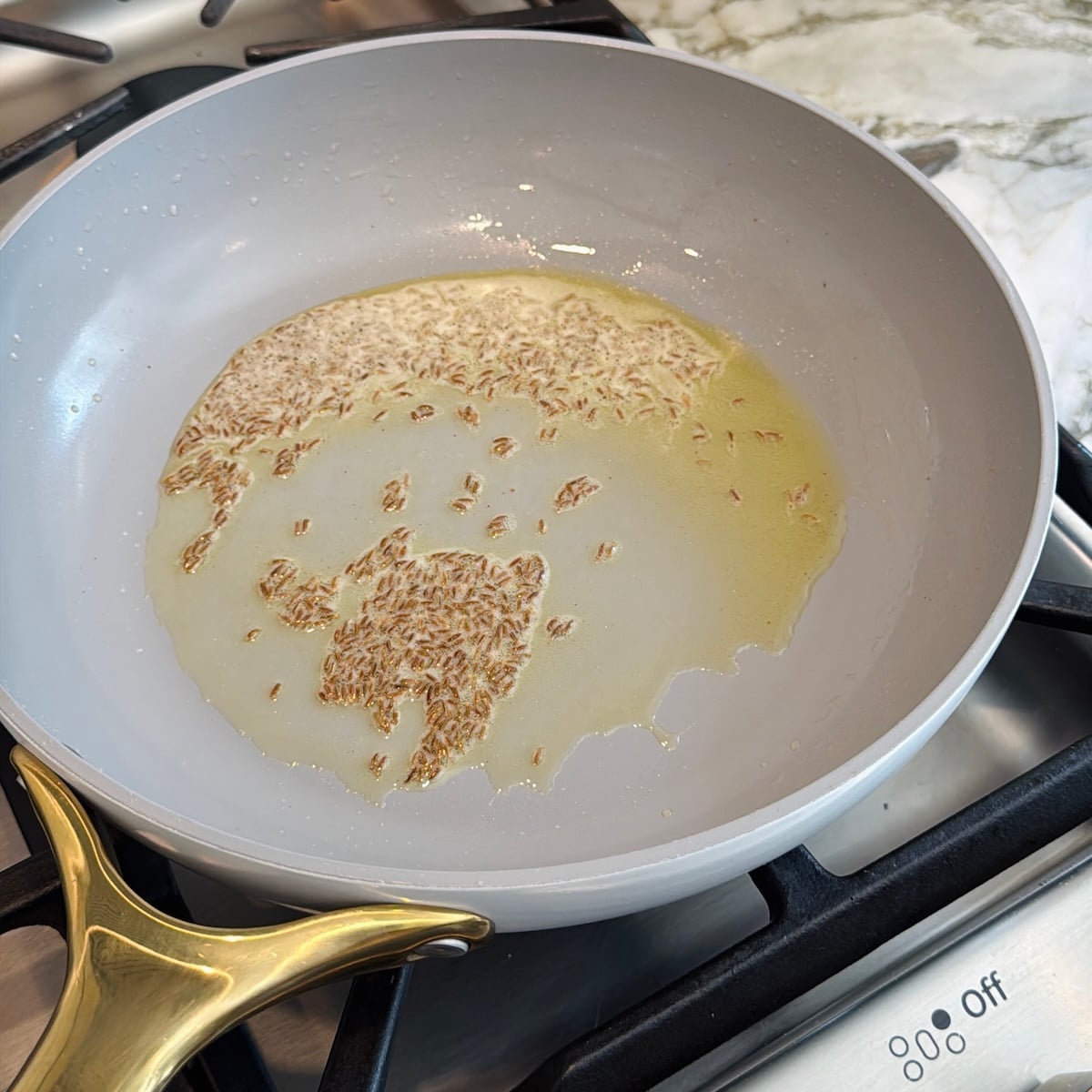 Cumin seeds frying in oil in a light gray pan on a gas stove, with a marble countertop visible in the background.