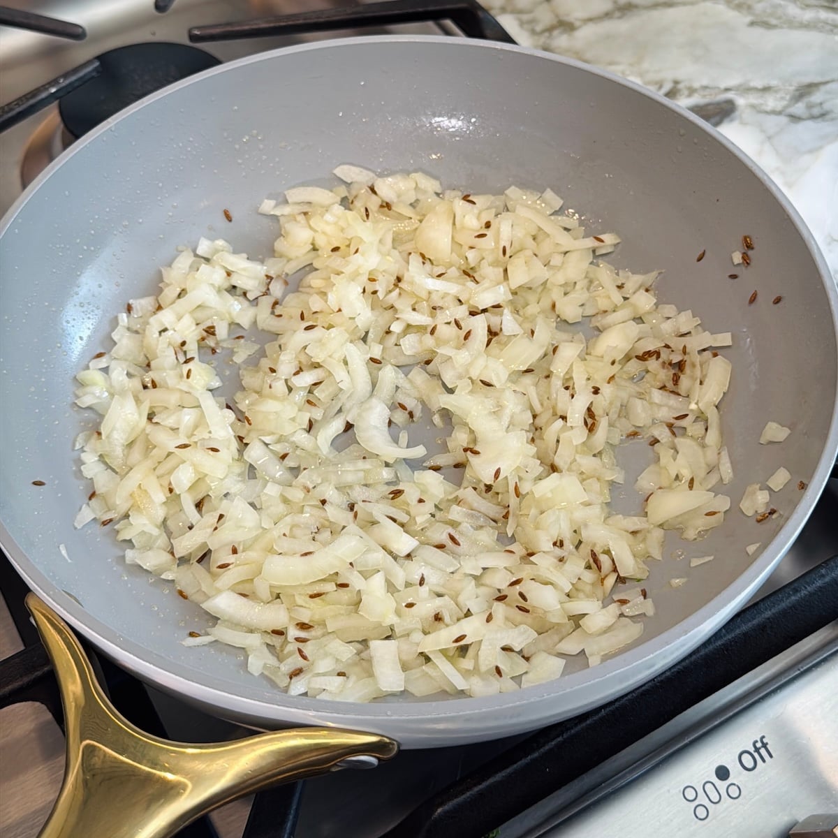 Chopped onions and cumin seeds sautéing in a gray frying pan on a stove.