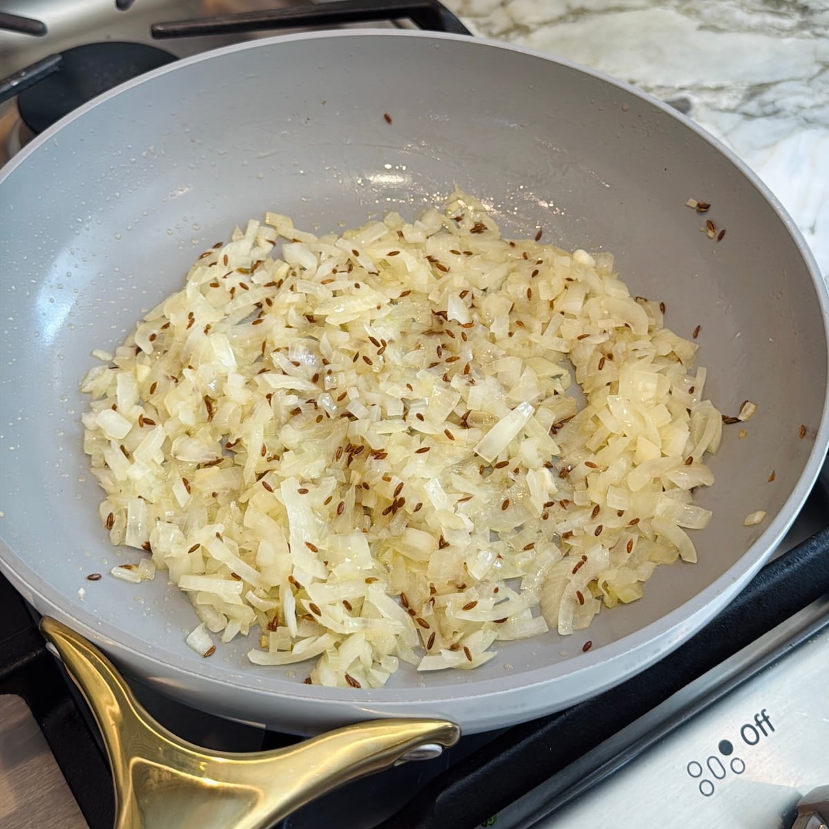 Chopped onions and cumin seeds are being sautéed in a light gray frying pan on a stovetop.