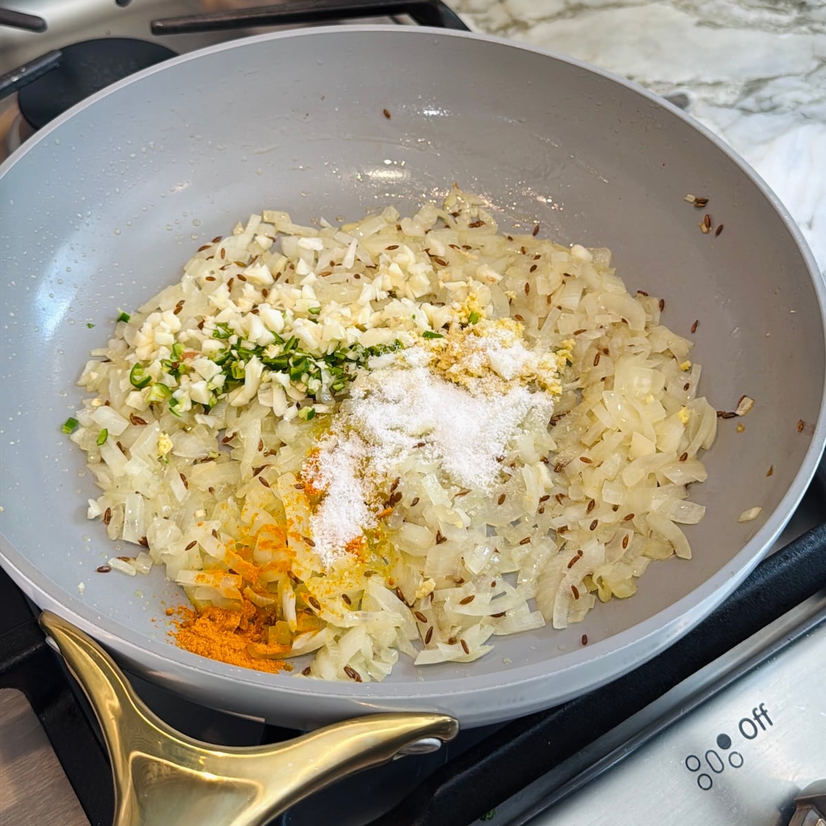 Chopped onions, garlic, green chili, ginger, salt, spices, and cumin seeds are being sautéed in a gray frying pan on a stovetop.
