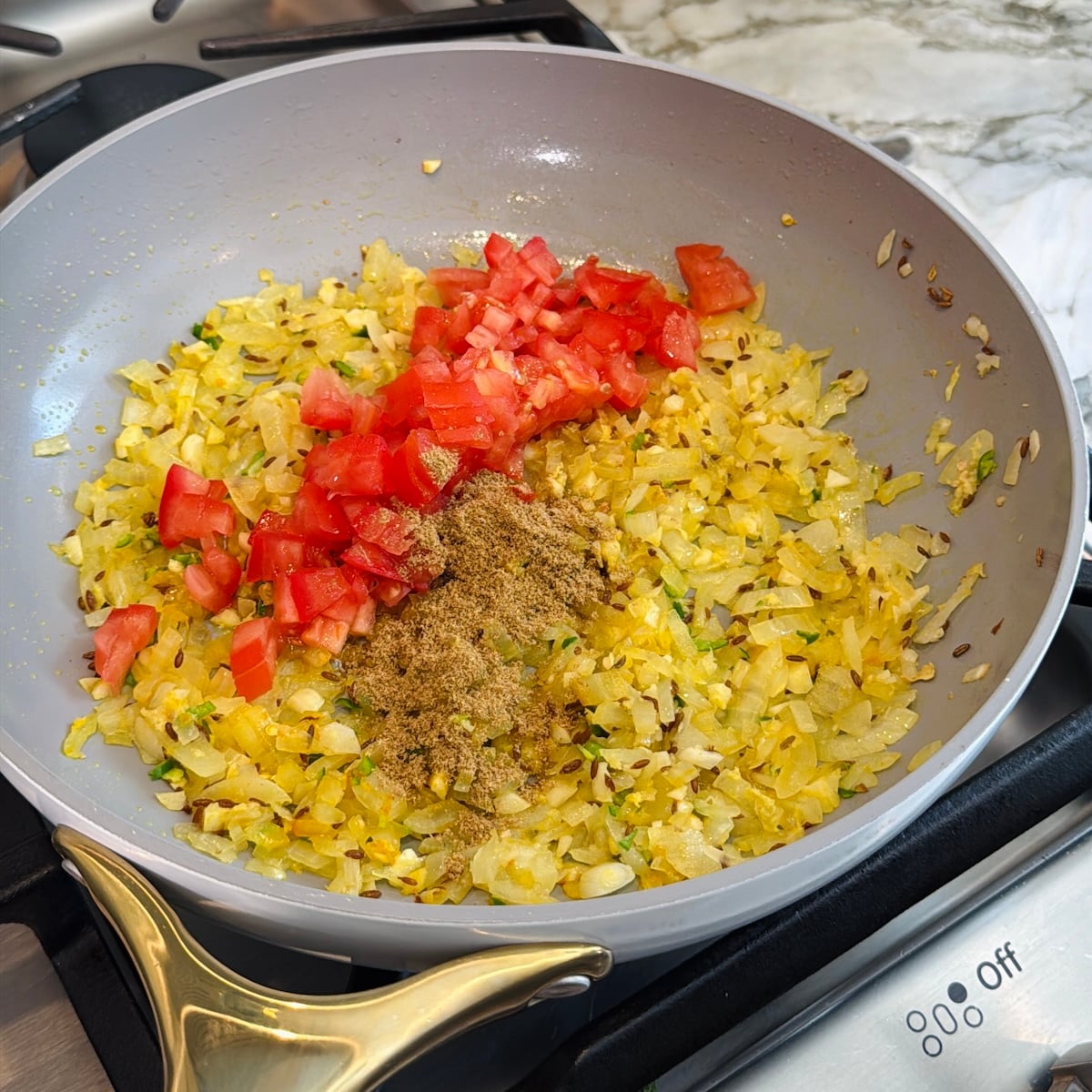 Chopped onions, tomatoes, and ground spices cooking in a frying pan on a stovetop.