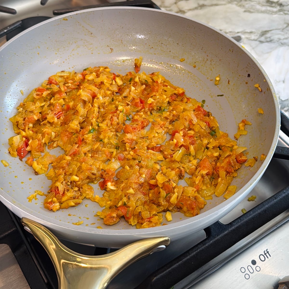 A frying pan on a stove contains sautéed onions, garlic, tomatoes, and spices.