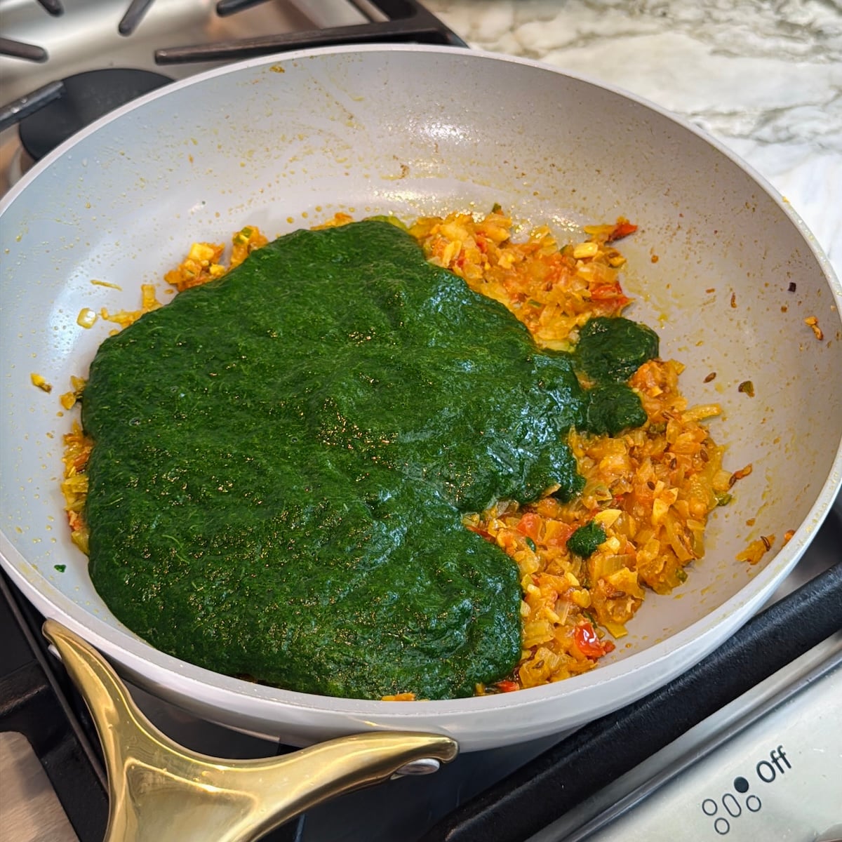 A pan on a stovetop contains sautéed onions and tomatoes with green spinach puree being added.