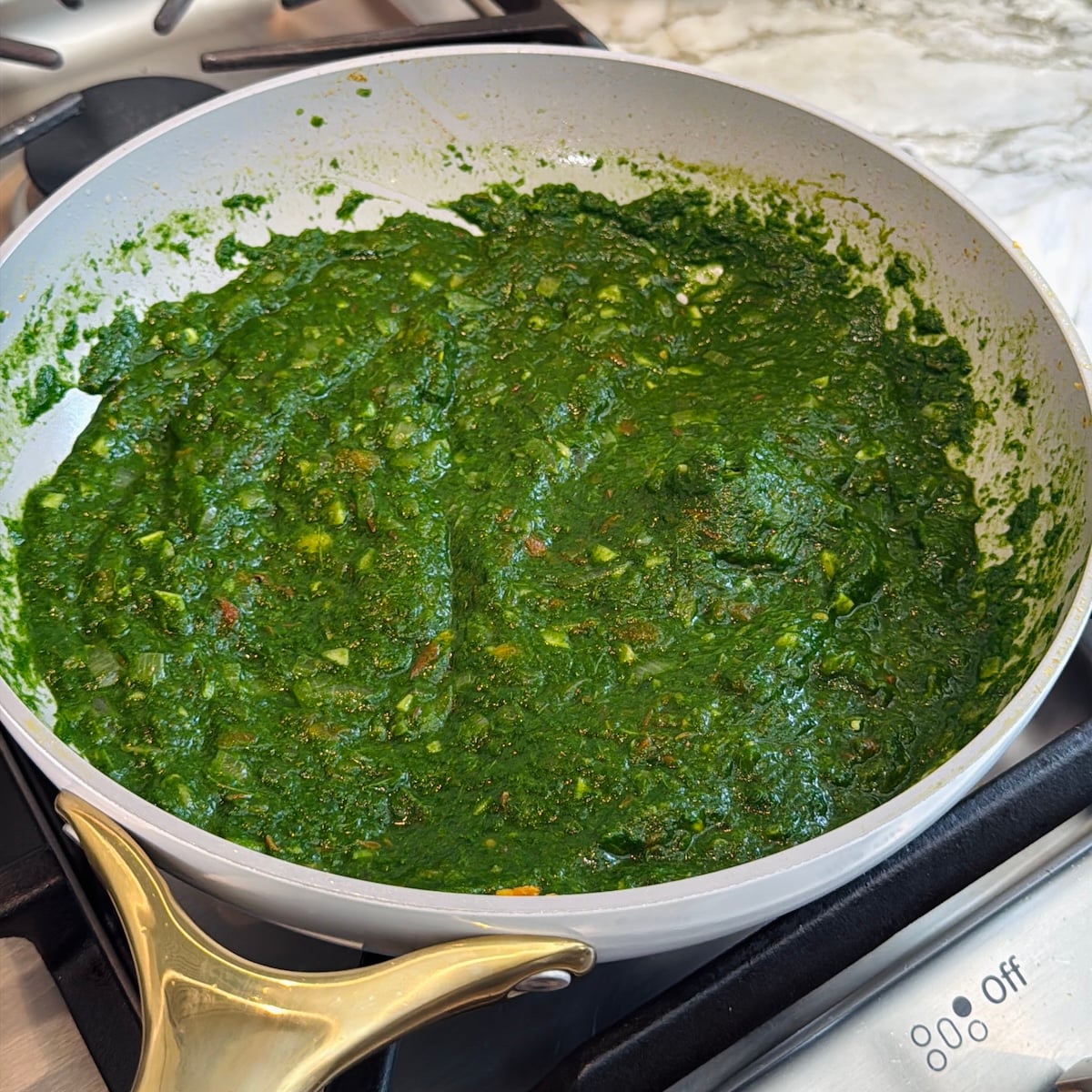 A pan filled with thick green spinach sauce cooking on a stove, with finely diced vegetables visible in the mixture.