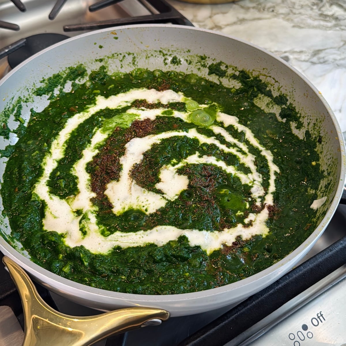 A pan of creamy spinach curry cooking on a stove, topped with a swirl of cream and sprinkled with spices.