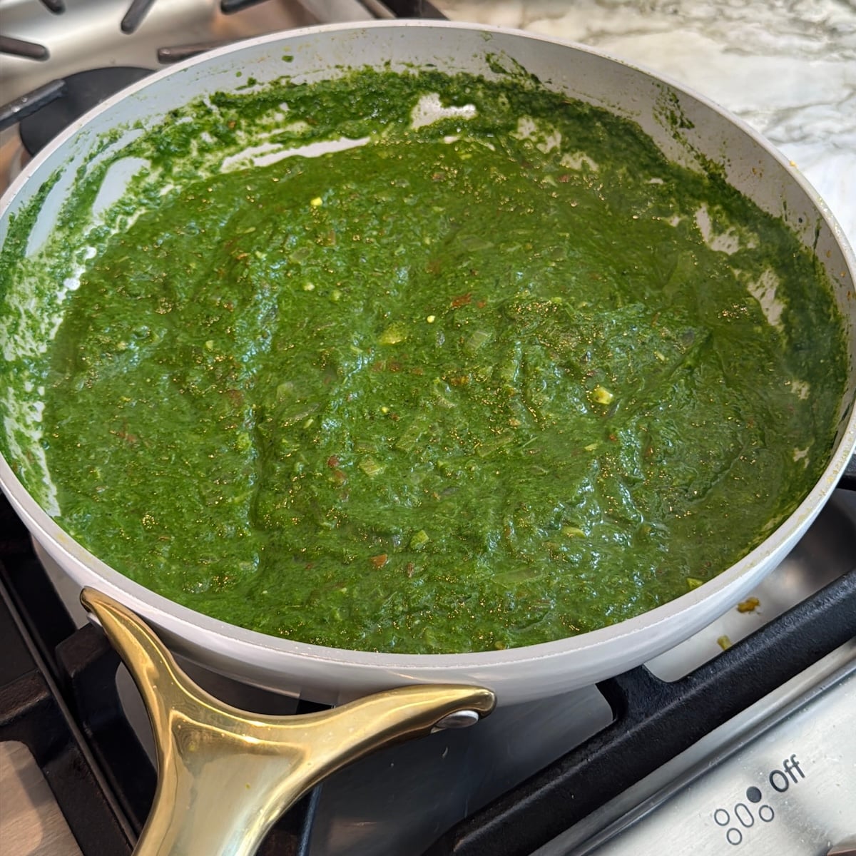 A white frying pan with a gold handle sits on a stove, filled with a thick, bright green spinach-based sauce or paste.
