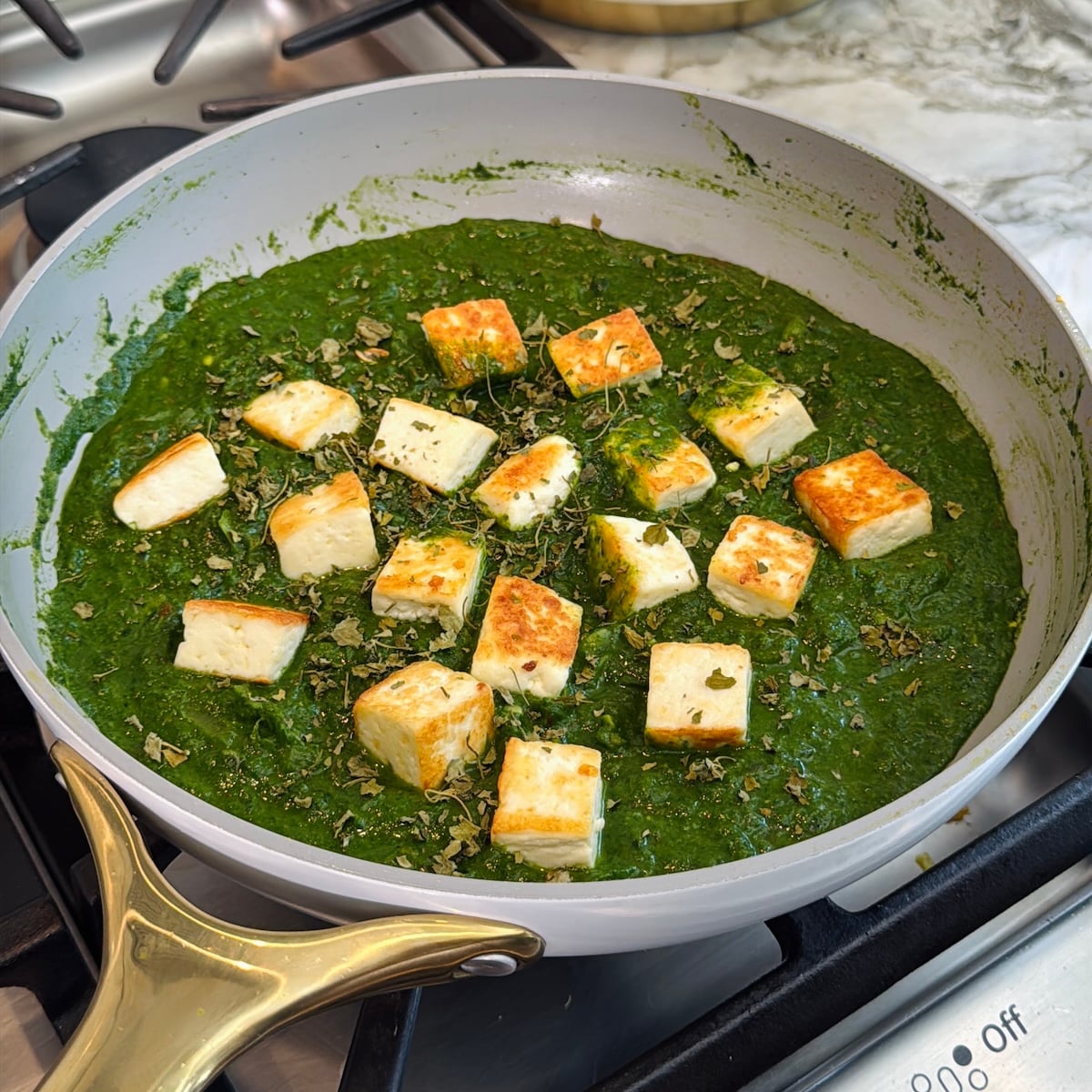 Cubes of paneer simmer in a green spinach sauce in a pan on a stovetop, representing the Indian dish palak paneer.