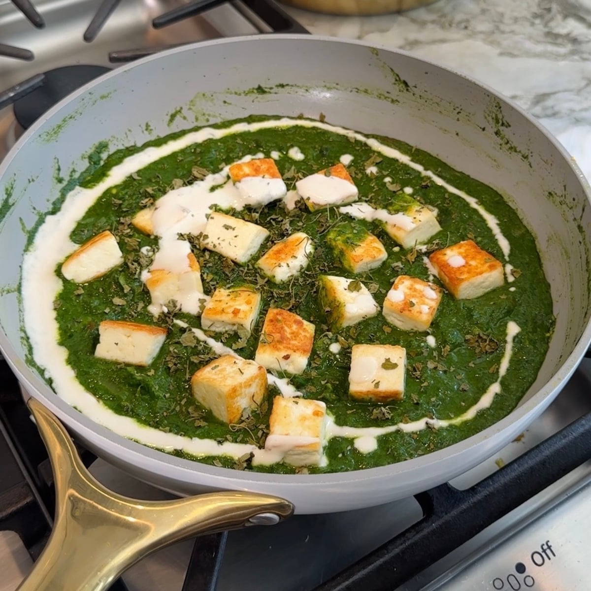 A pan on a stove contains palak paneer, with cubes of paneer in green spinach gravy, garnished with cream and herbs.