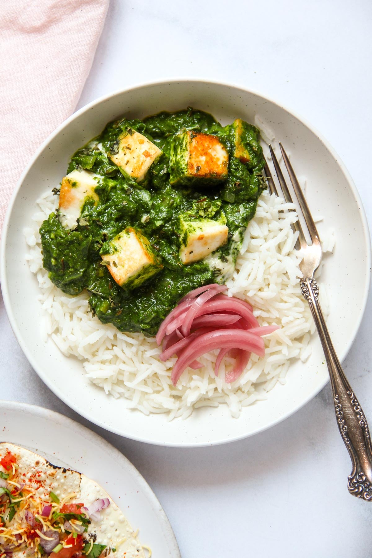 A white plate with white rice topped with spinach curry and paneer cubes, garnished with pickled red onions. A fork rests on the side of the plate.