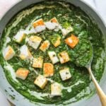 A skillet of palak paneer, featuring cubes of paneer cheese in a creamy spinach sauce, garnished with herbs and cream, with a serving spoon on the side.