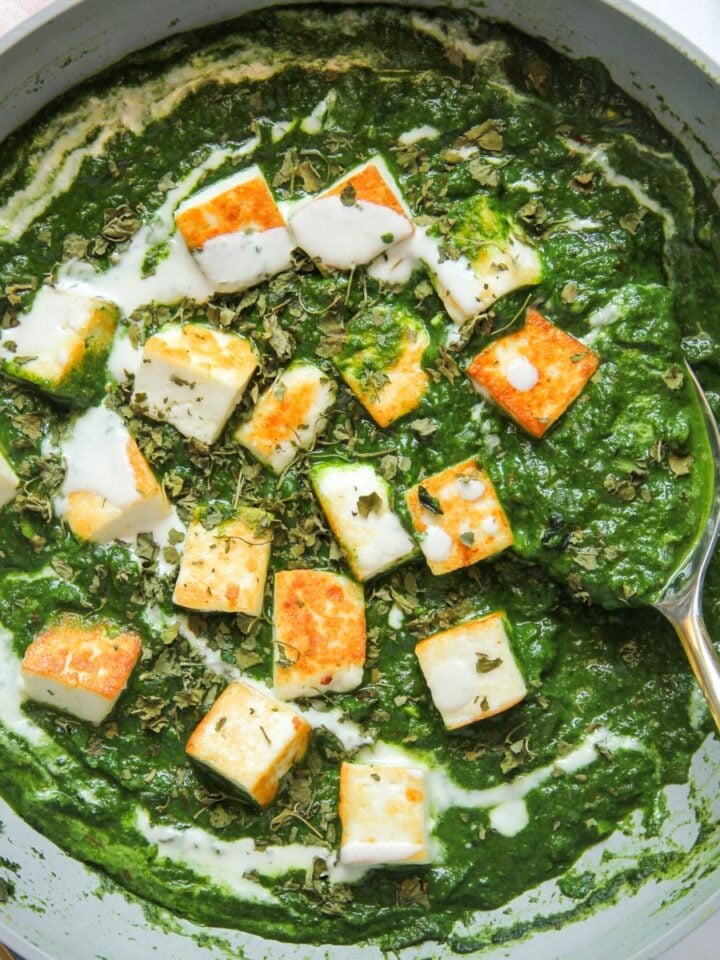 A skillet of palak paneer, featuring cubes of paneer cheese in a creamy spinach sauce, garnished with herbs and cream, with a serving spoon on the side.