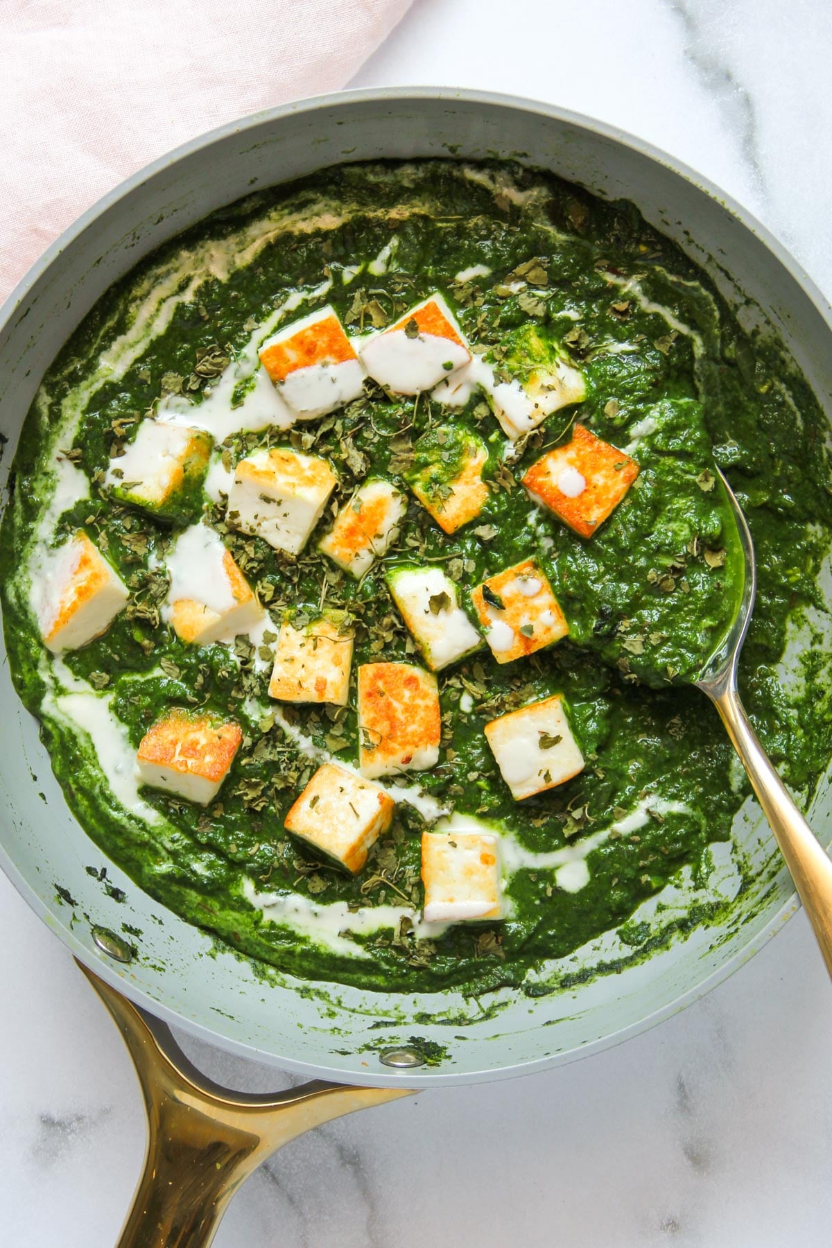 A pan of palak paneer with golden paneer cubes in creamy spinach sauce, garnished with herbs, and a spoon resting inside.