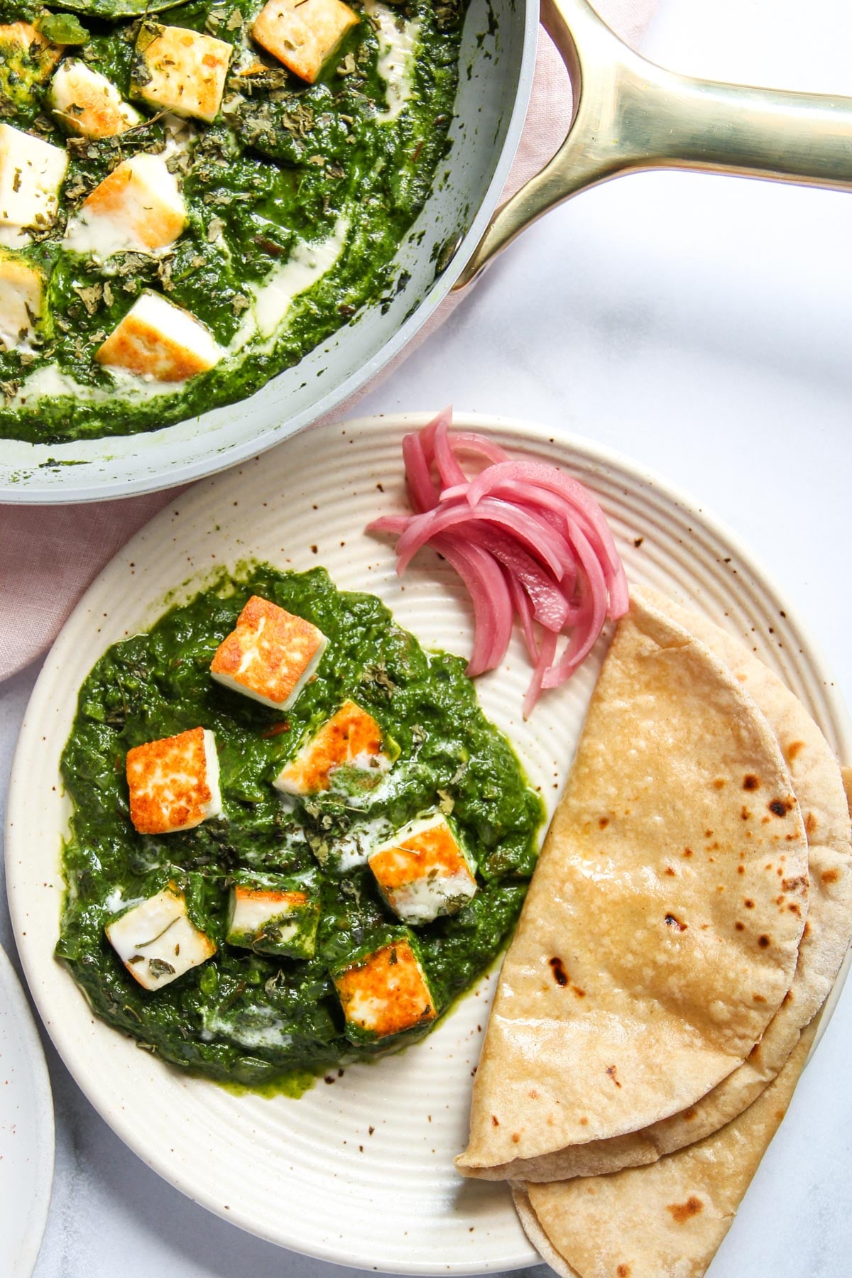 A plate with palak paneer, two rotis, and pickled onions next to a pan containing more palak paneer.