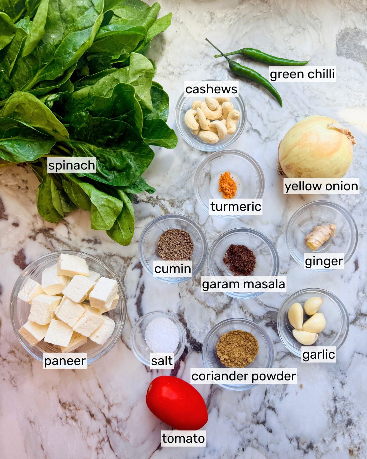 Ingredients for a spinach paneer dish, including spinach, paneer, tomato, spices, garlic, ginger, onion, green chilli, and cashews on a marble surface.