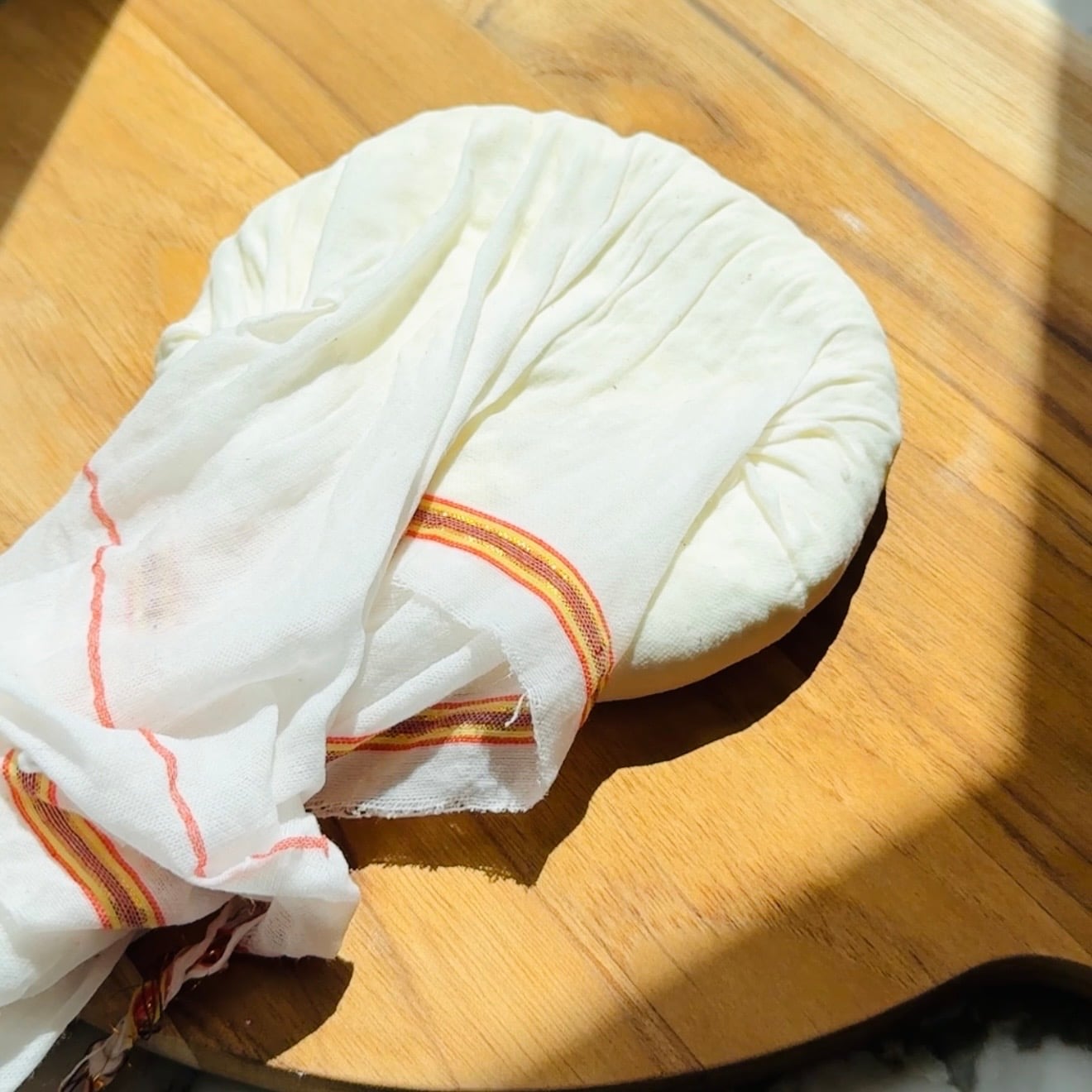 Round ball of white cheese wrapped in a cloth with a colored edge, resting on a wooden surface in sunlight.