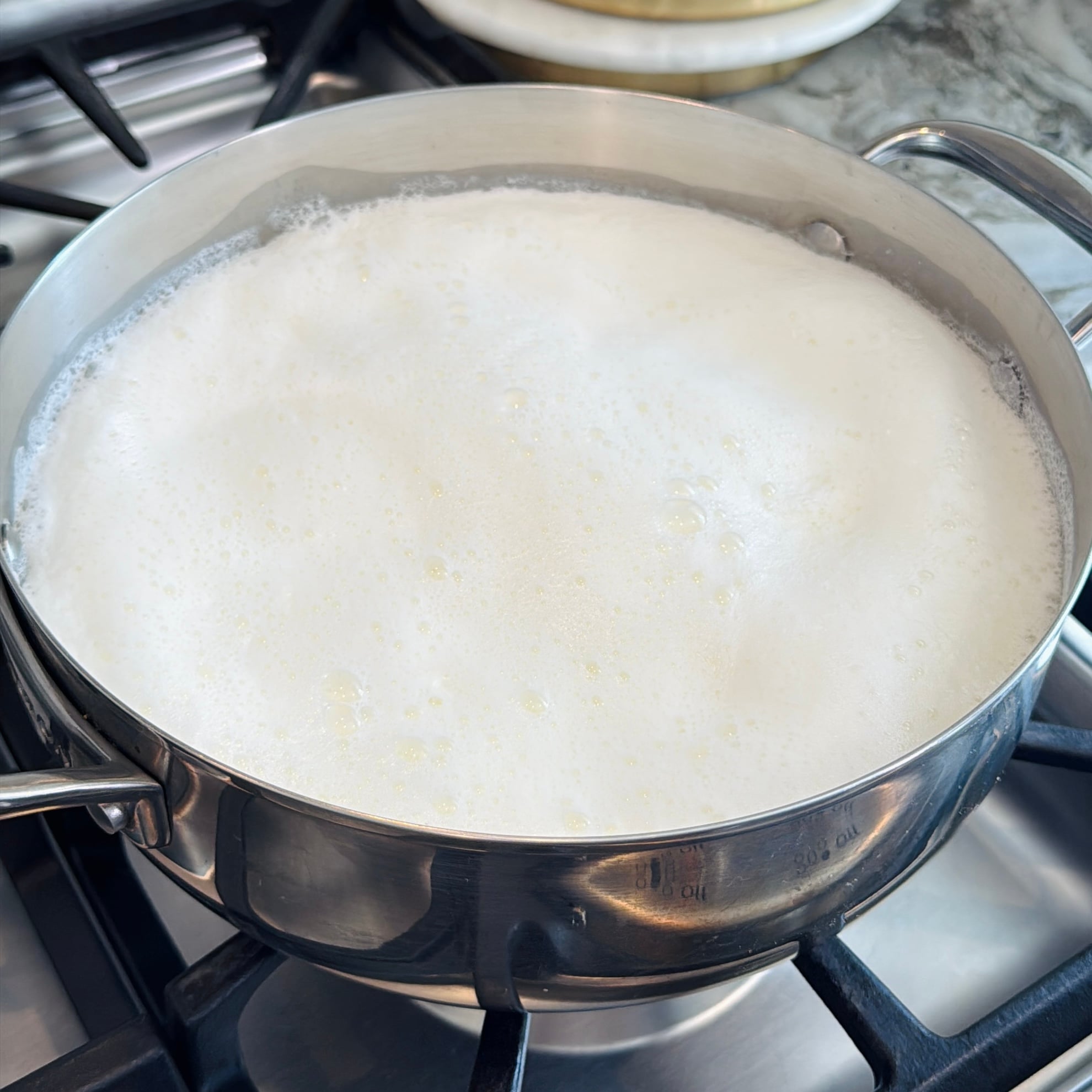 A stainless steel pot of milk is boiling on a stove, with foam rising to the top and small bubbles visible.