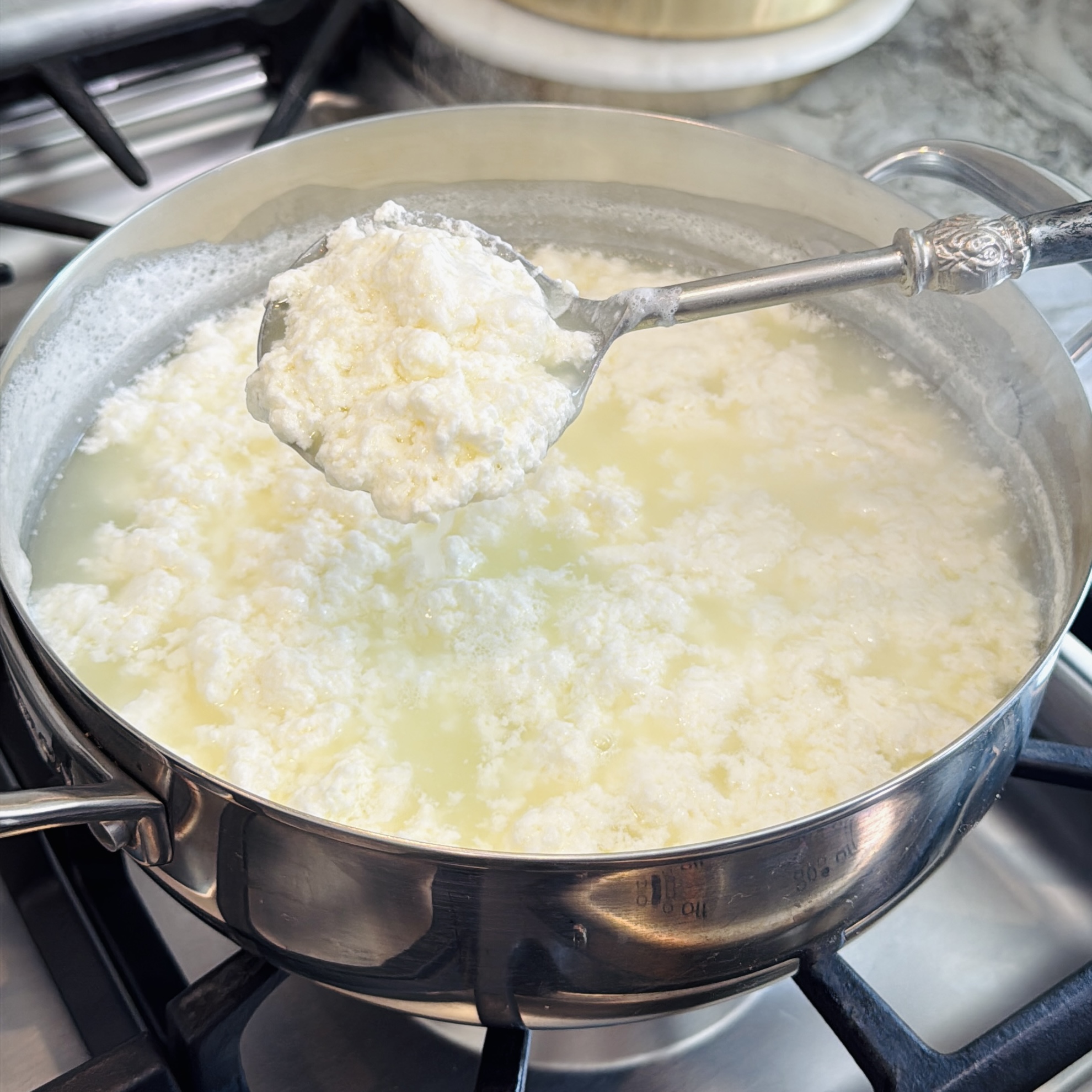 A slotted spoon lifts fresh cheese curds from a pot of hot whey on a stovetop during the cheese-making process.