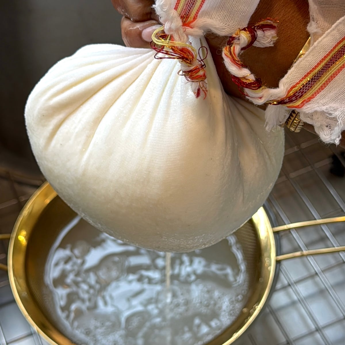 A hand holds a cloth bundle of fresh cheese over a pot, draining whey through the fabric.