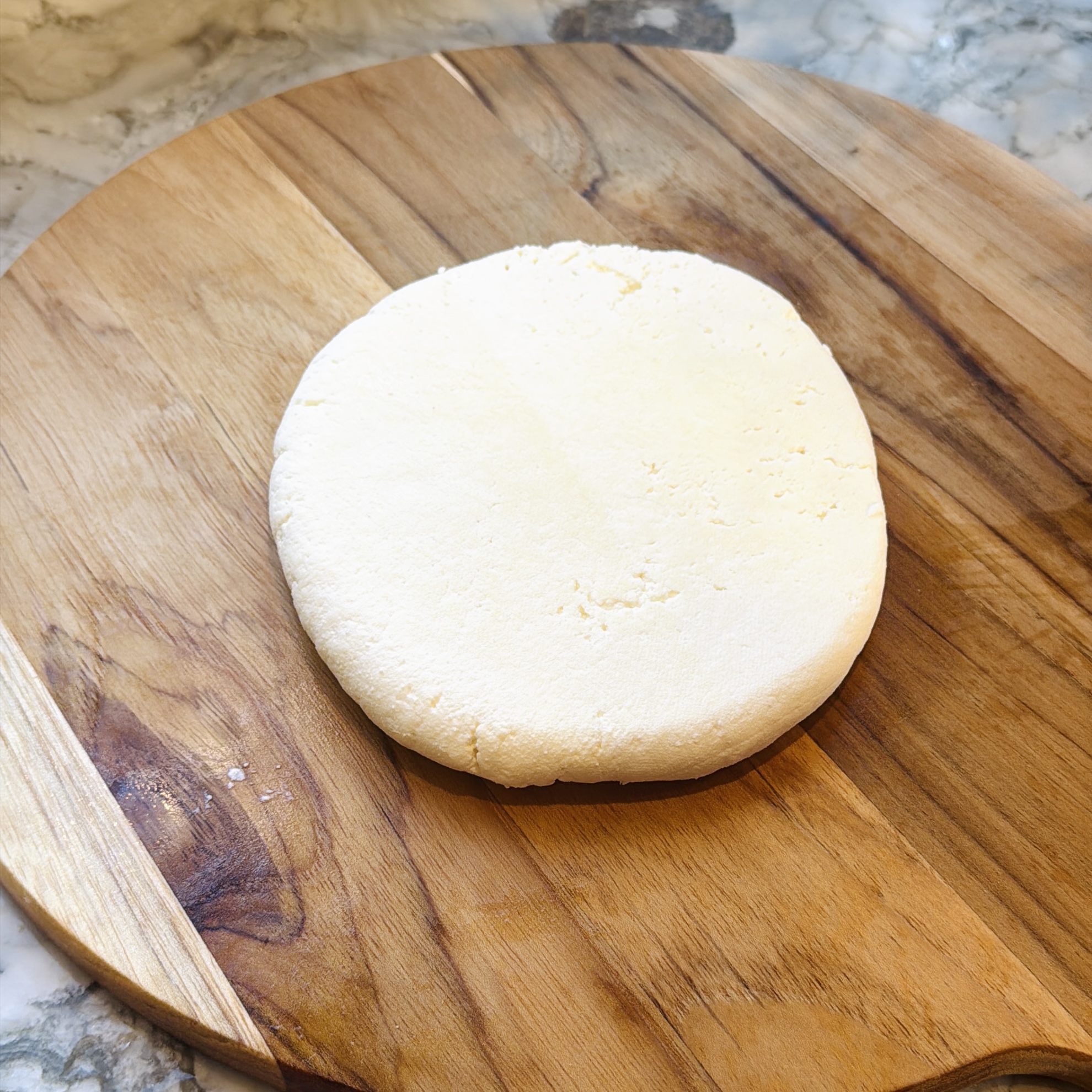 A round, flattened disk of fresh white cheese sits on a wooden cutting board, with a marble countertop in the background.