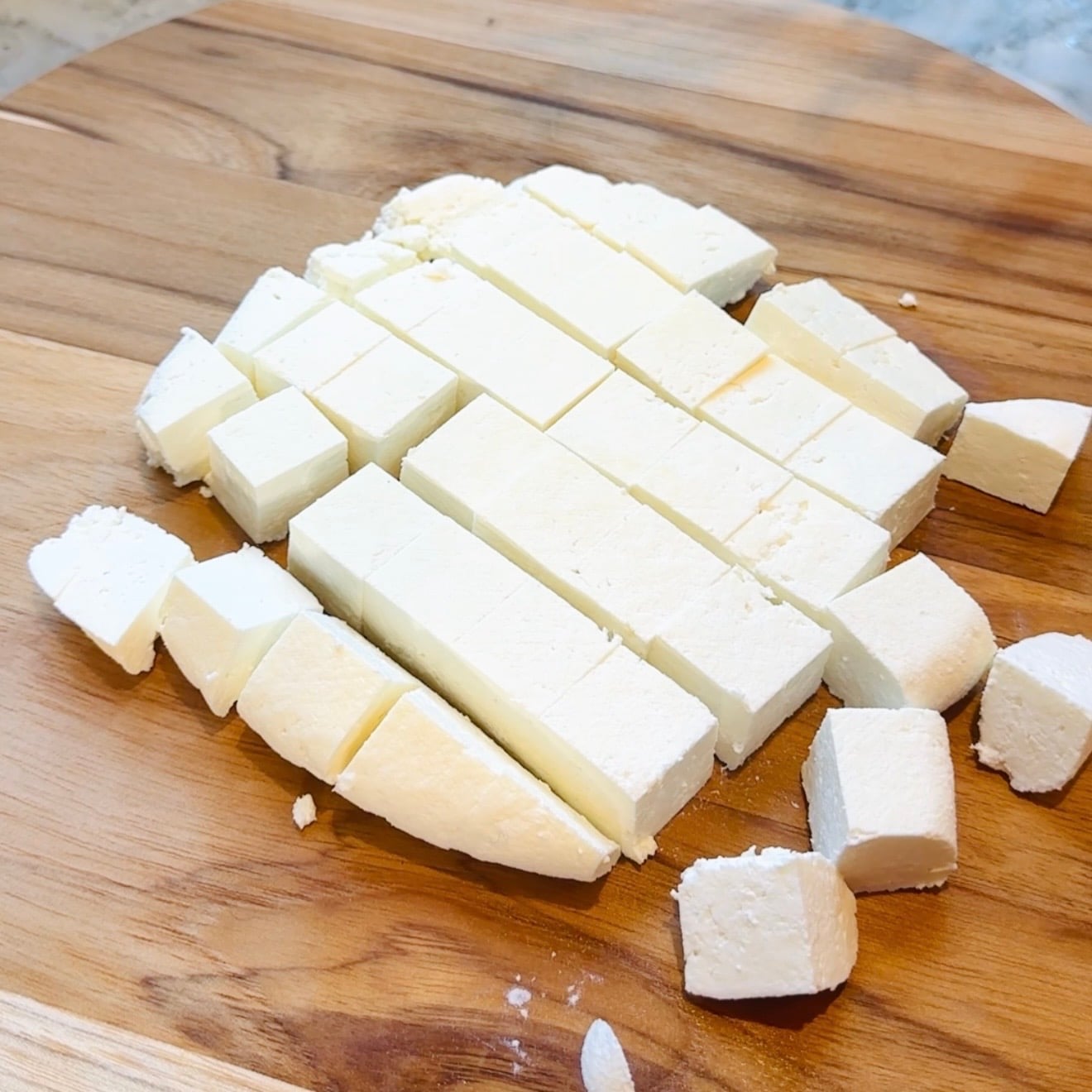 Blocks of white cheese cut into even rectangular and cube pieces are arranged on a wooden cutting board.