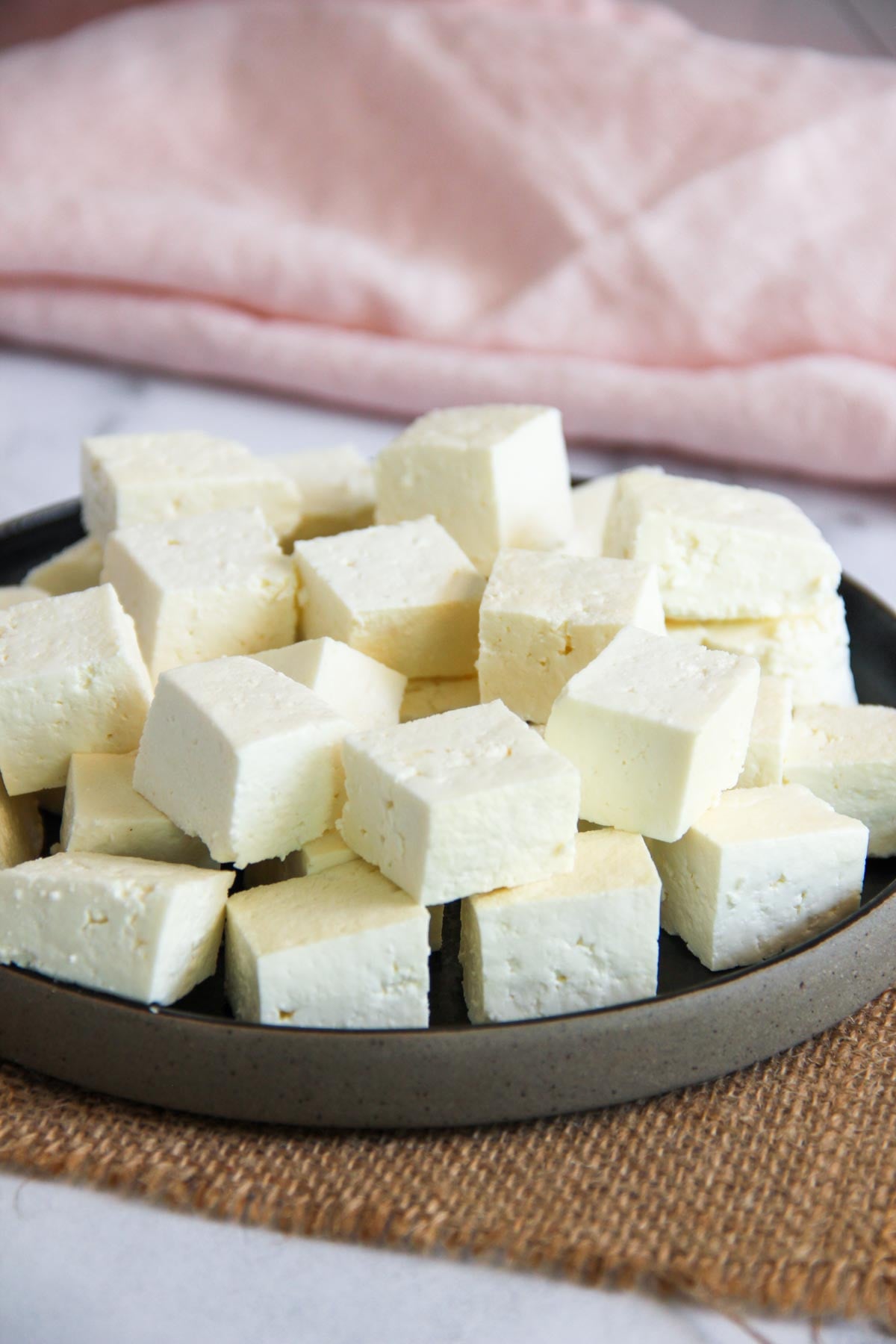 A plate filled with cubed pieces of paneer cheese sits on a woven mat, with a pink cloth in the background.