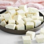 A plate filled with neatly arranged cubes of white paneer cheese, with a few pieces scattered on the white surface next to the plate.