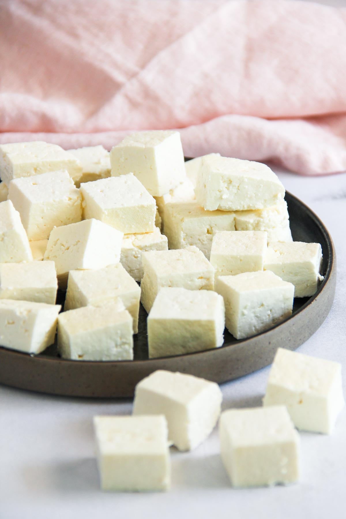 A plate of cubed paneer cheese is arranged on a dark round dish, with a few cubes scattered in the foreground and a light pink cloth in the background.