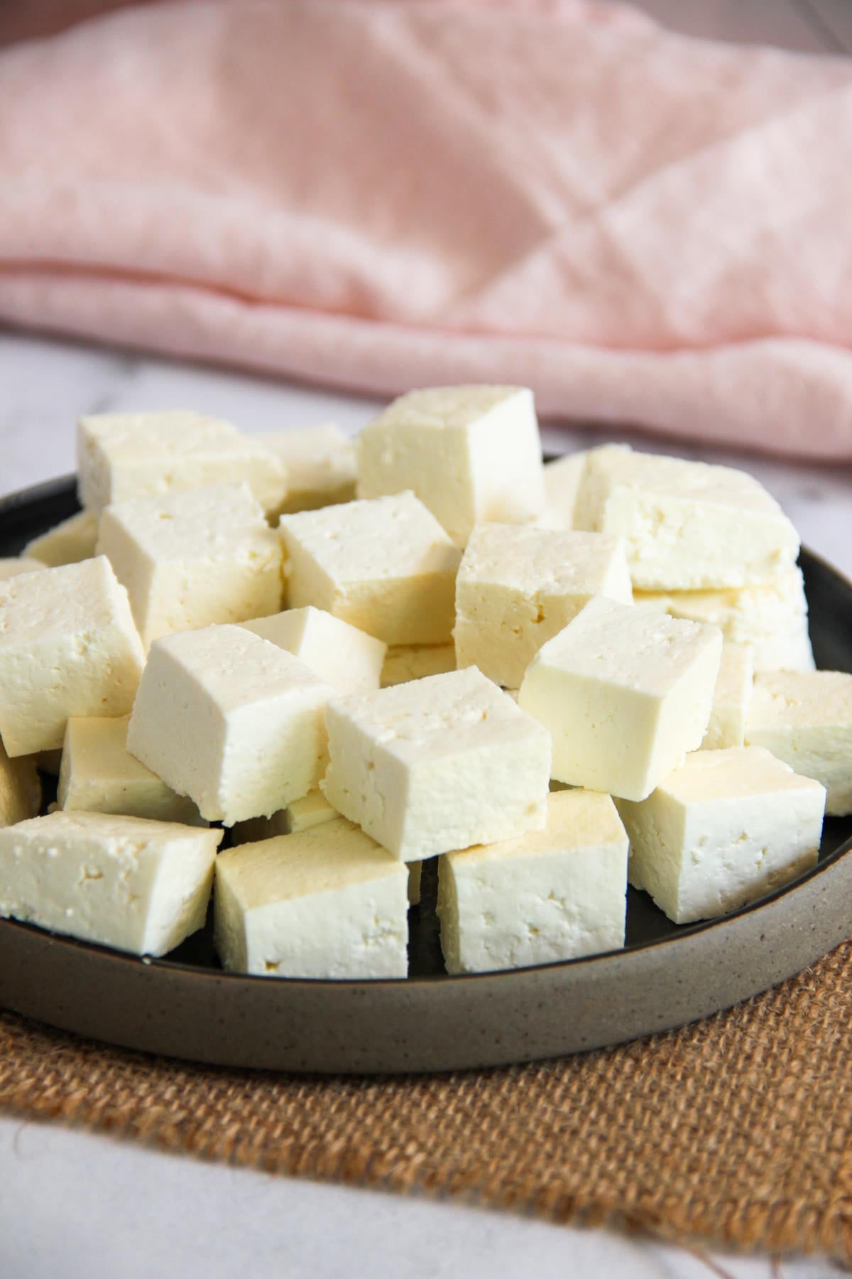 A black plate holds several cubes of white paneer cheese, arranged in a pile. A pink cloth and brown mat are in the background.