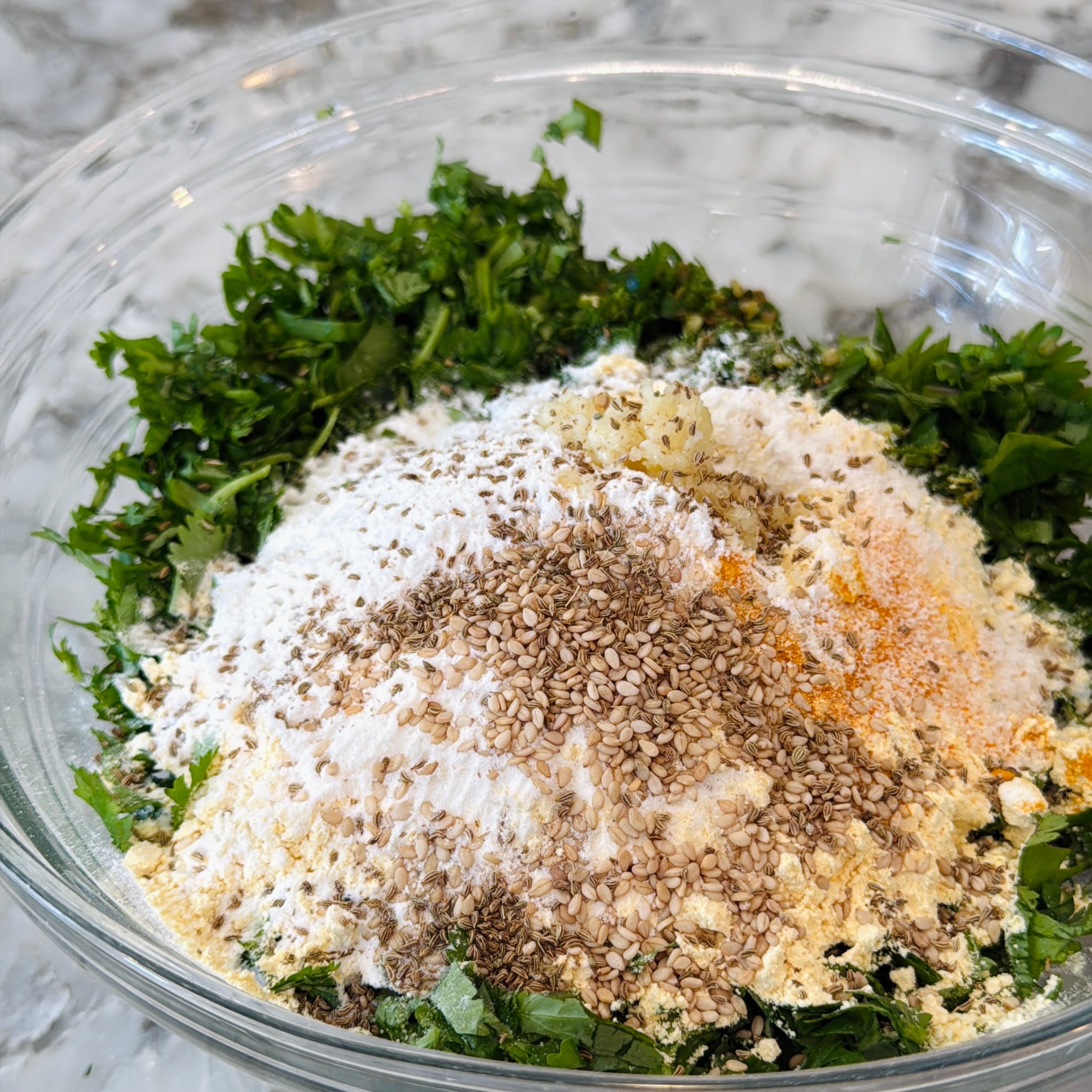 A glass bowl containing chopped herbs, flour, sesame seeds, grated garlic, and various spices on a marble countertop.