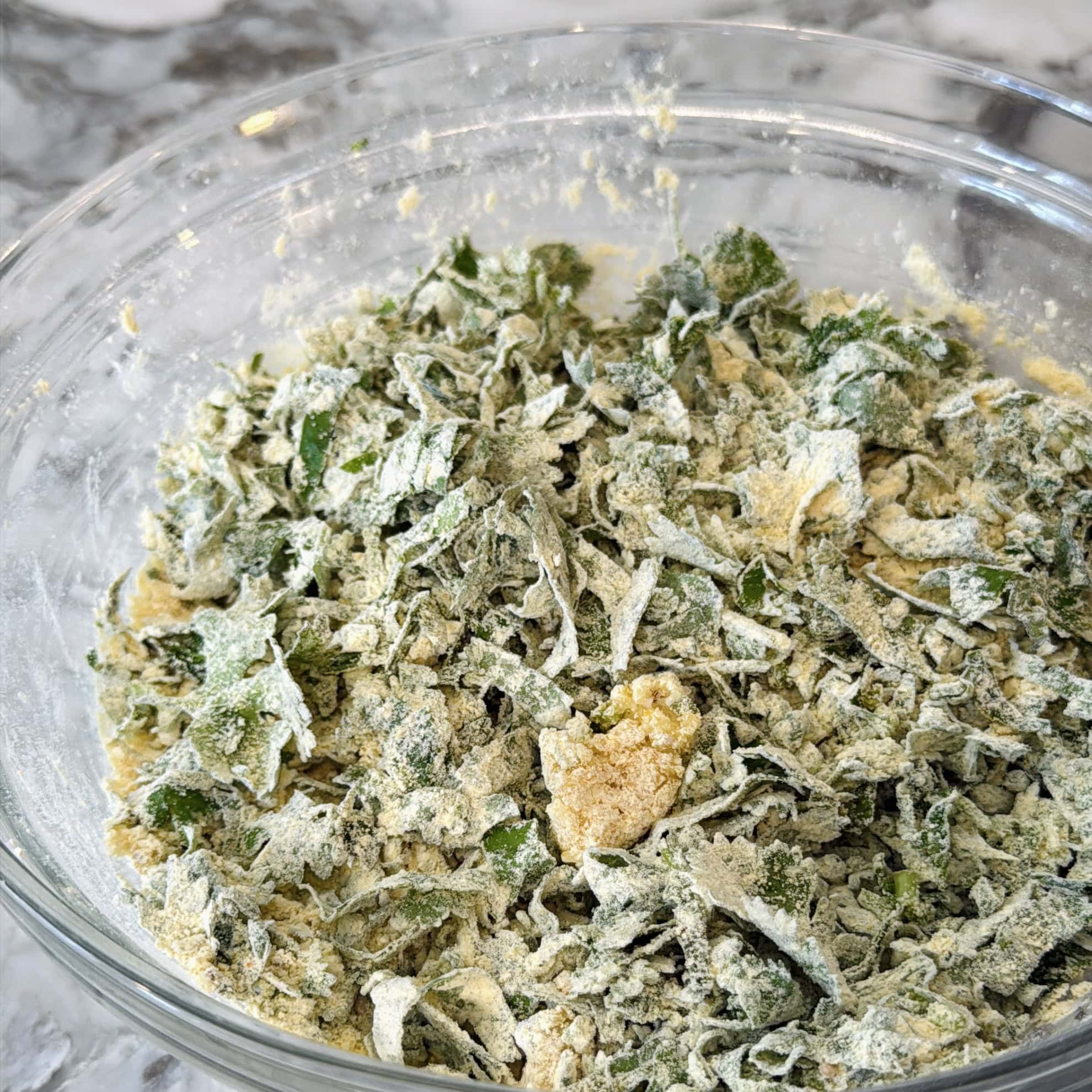 A clear glass bowl filled with chopped leafy greens coated in a yellow flour mixture, set on a marble countertop.
