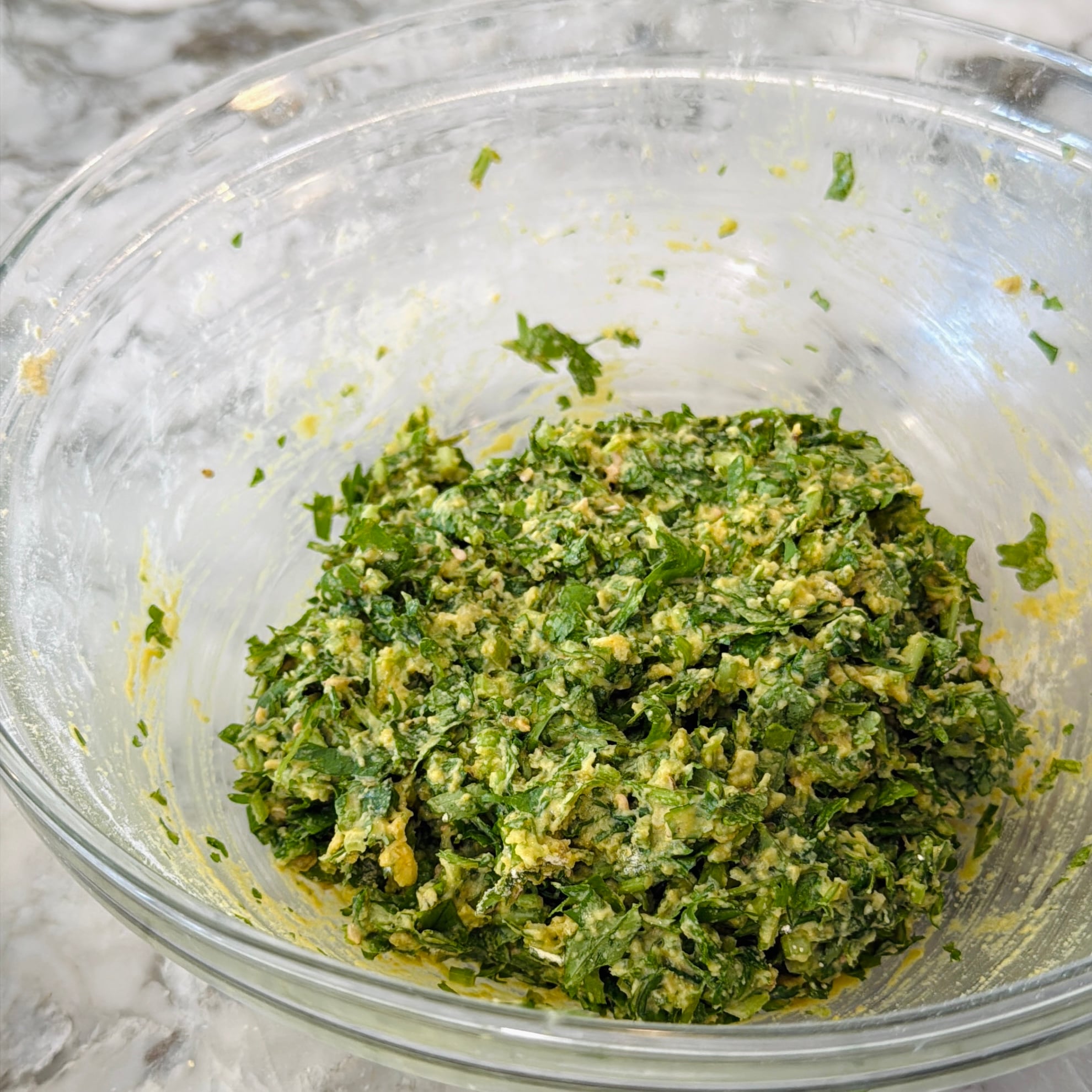 A glass bowl containing a mixture of chopped green herbs and a yellow batter on a marble countertop.