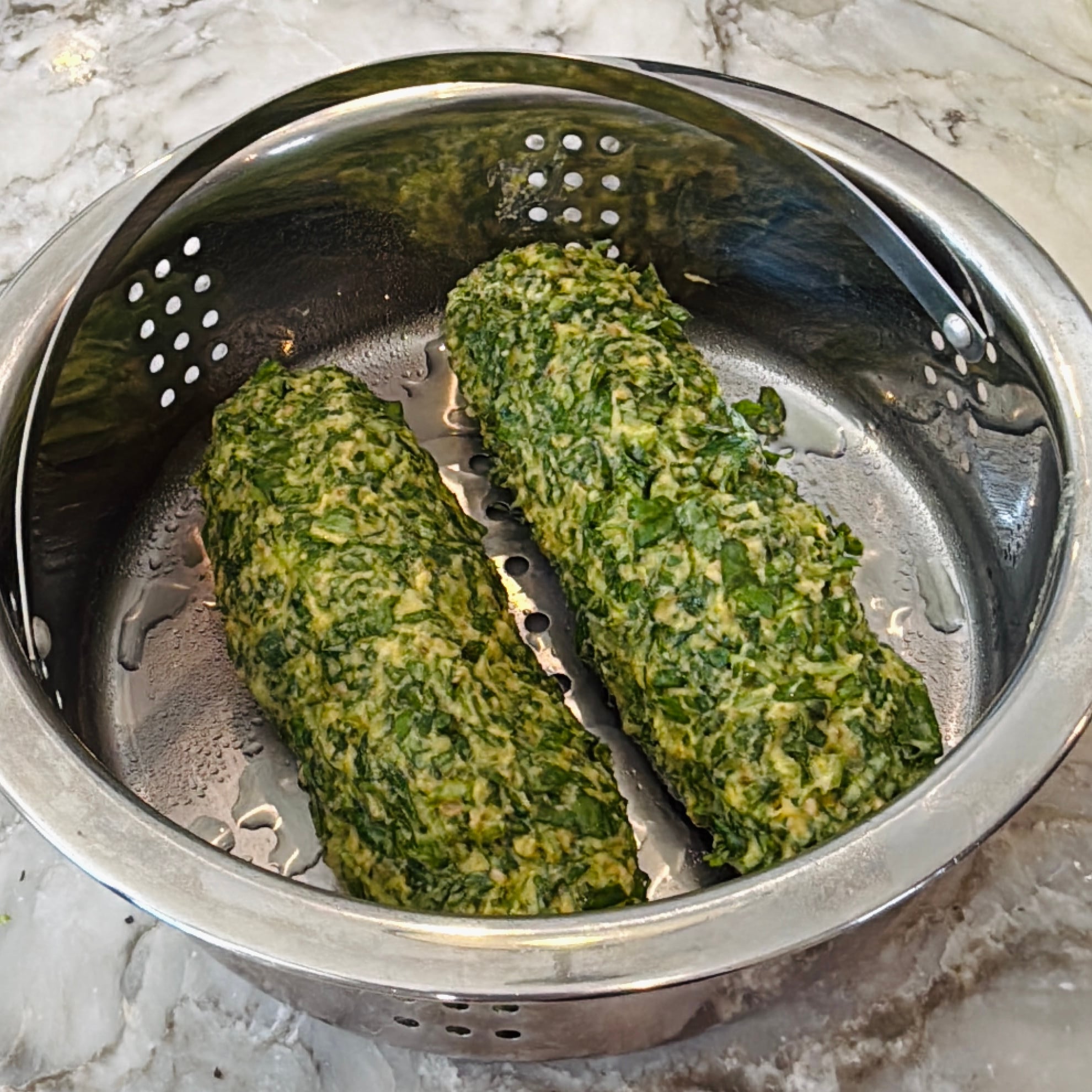 Two logs of uncooked green spinach mixture rest in a metal steamer basket on a marble countertop.