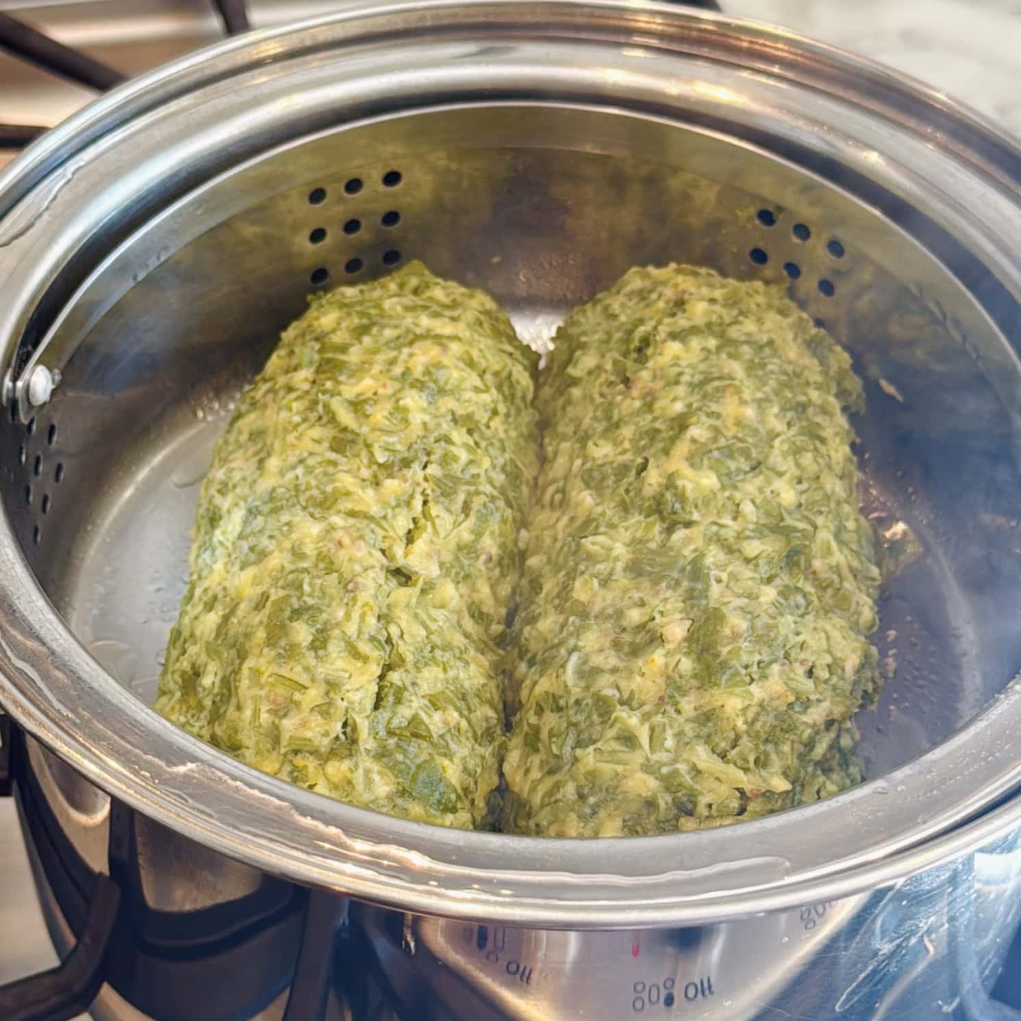 Two green vegetable loaves are steaming in a metal steamer basket on a stovetop. Steam is visible rising from the pot.