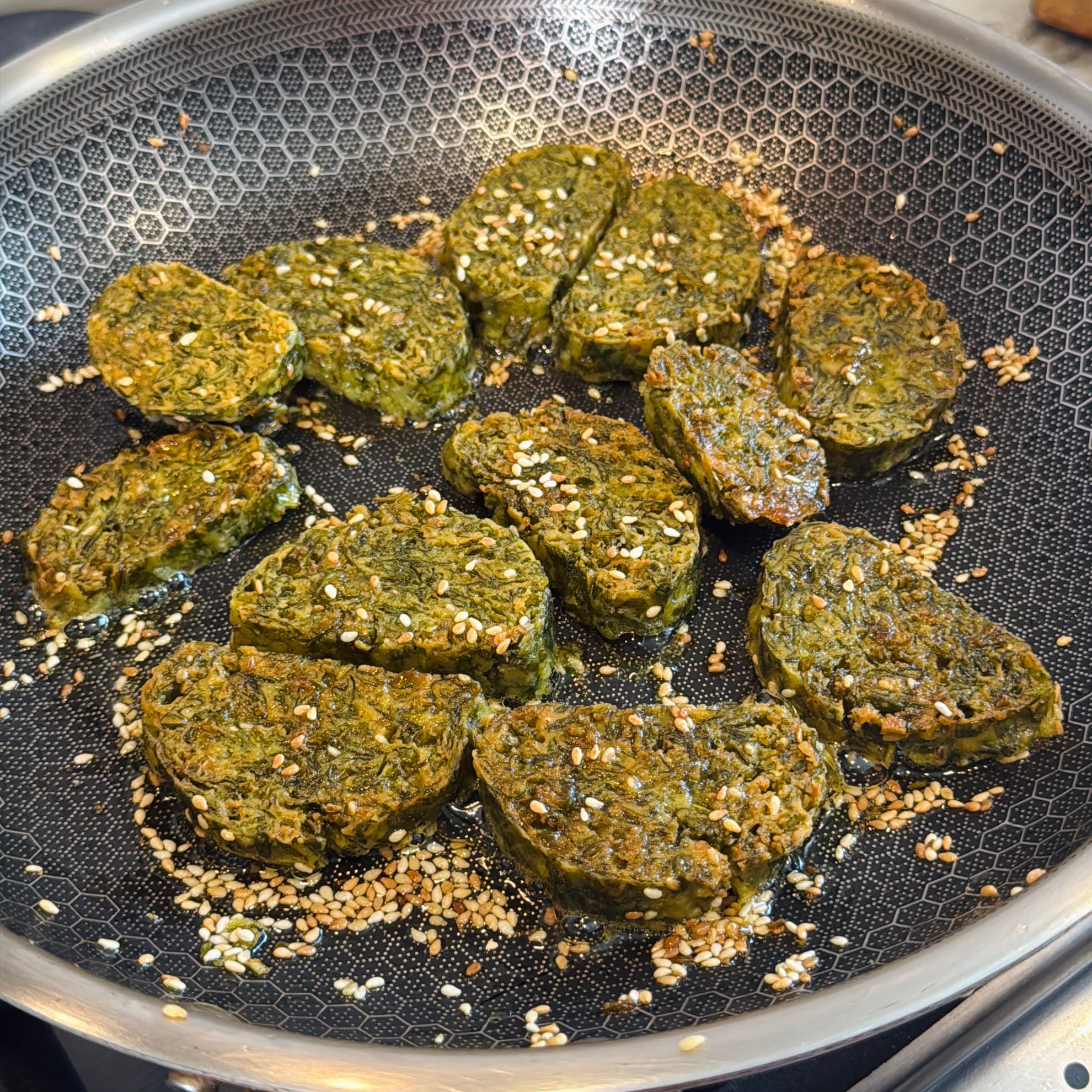Sliced pieces of pan-fried green vegetable patties with sesame seeds cooking in a nonstick frying pan.