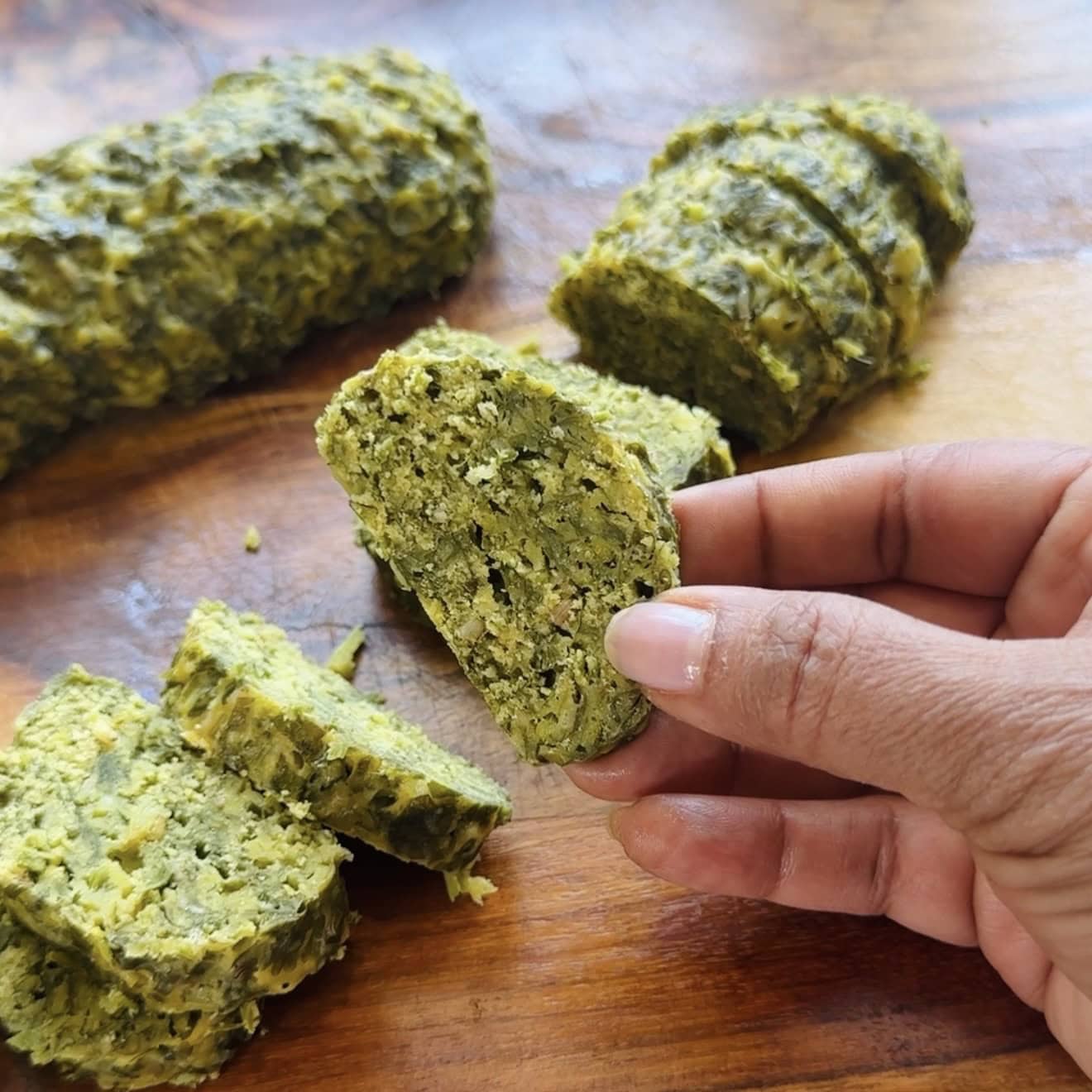 A hand holding a slice of green, herb-filled log-shaped food, with more slices and an unsliced portion on a wooden surface in the background.