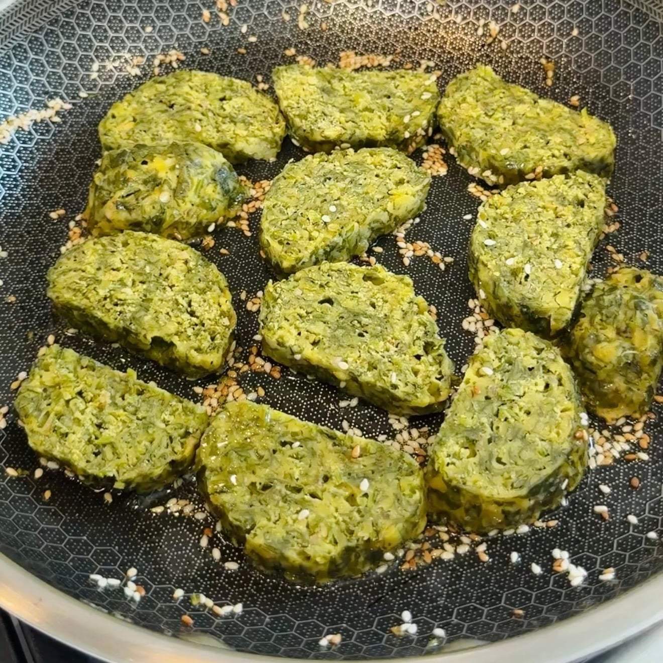 Several green falafel patties are cooking in a nonstick frying pan, sprinkled with sesame seeds.