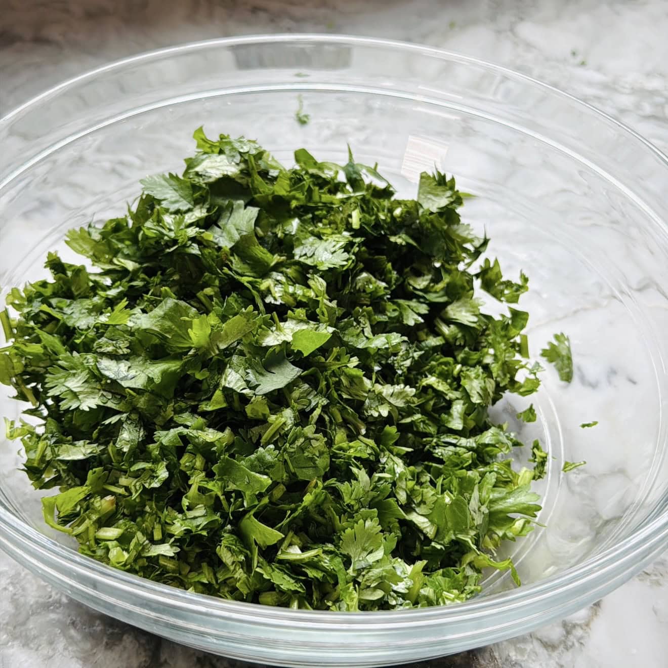 A clear glass bowl filled with freshly chopped cilantro on a marble surface.