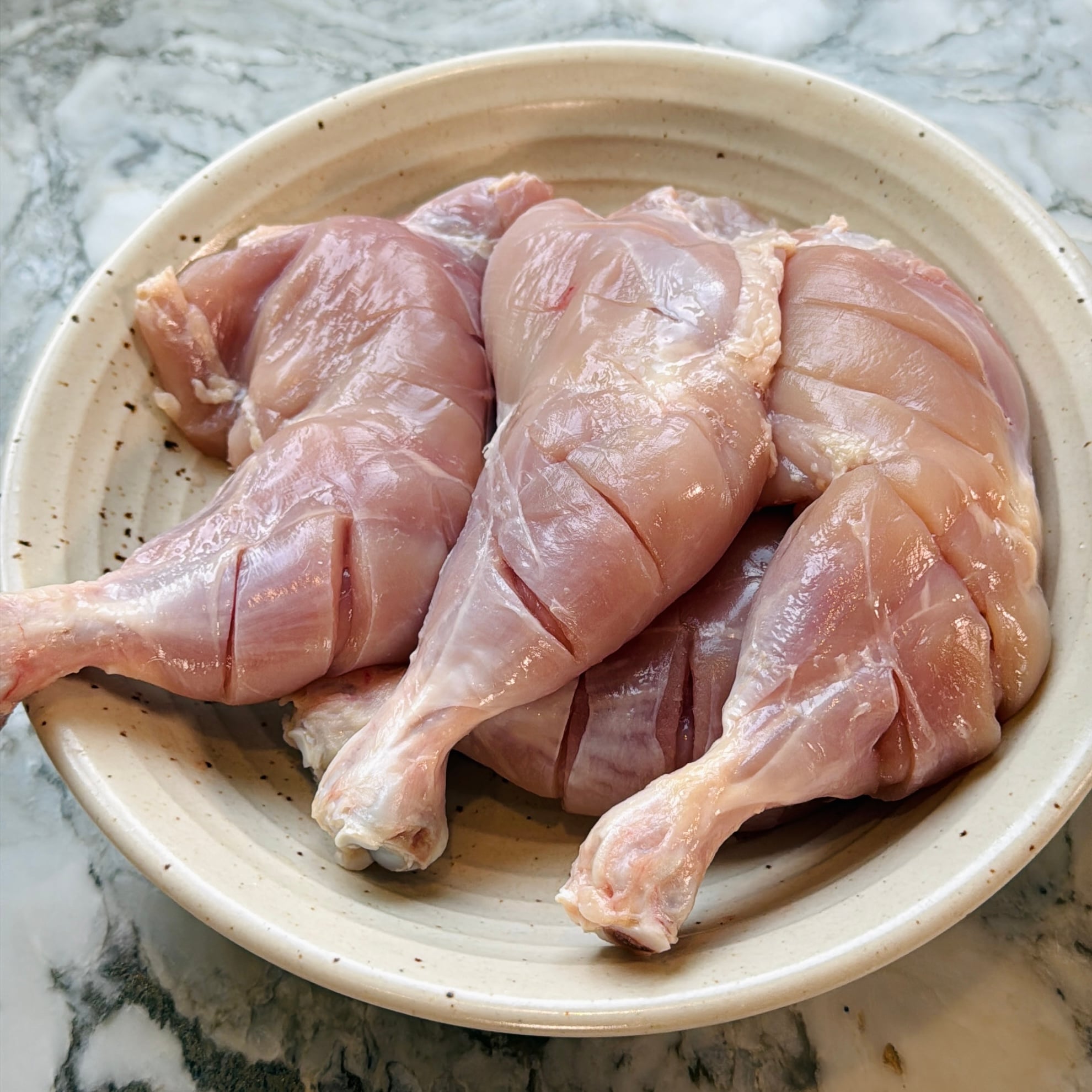 Four raw, skinless chicken legs with shallow cuts placed in a beige ceramic bowl on a marble countertop.