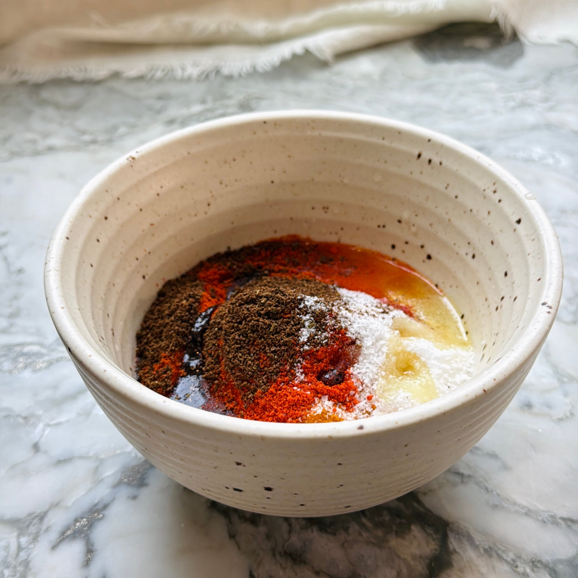 A ceramic bowl on a marble surface containing various ground spices, salt, oil, and yogurt before mixing.