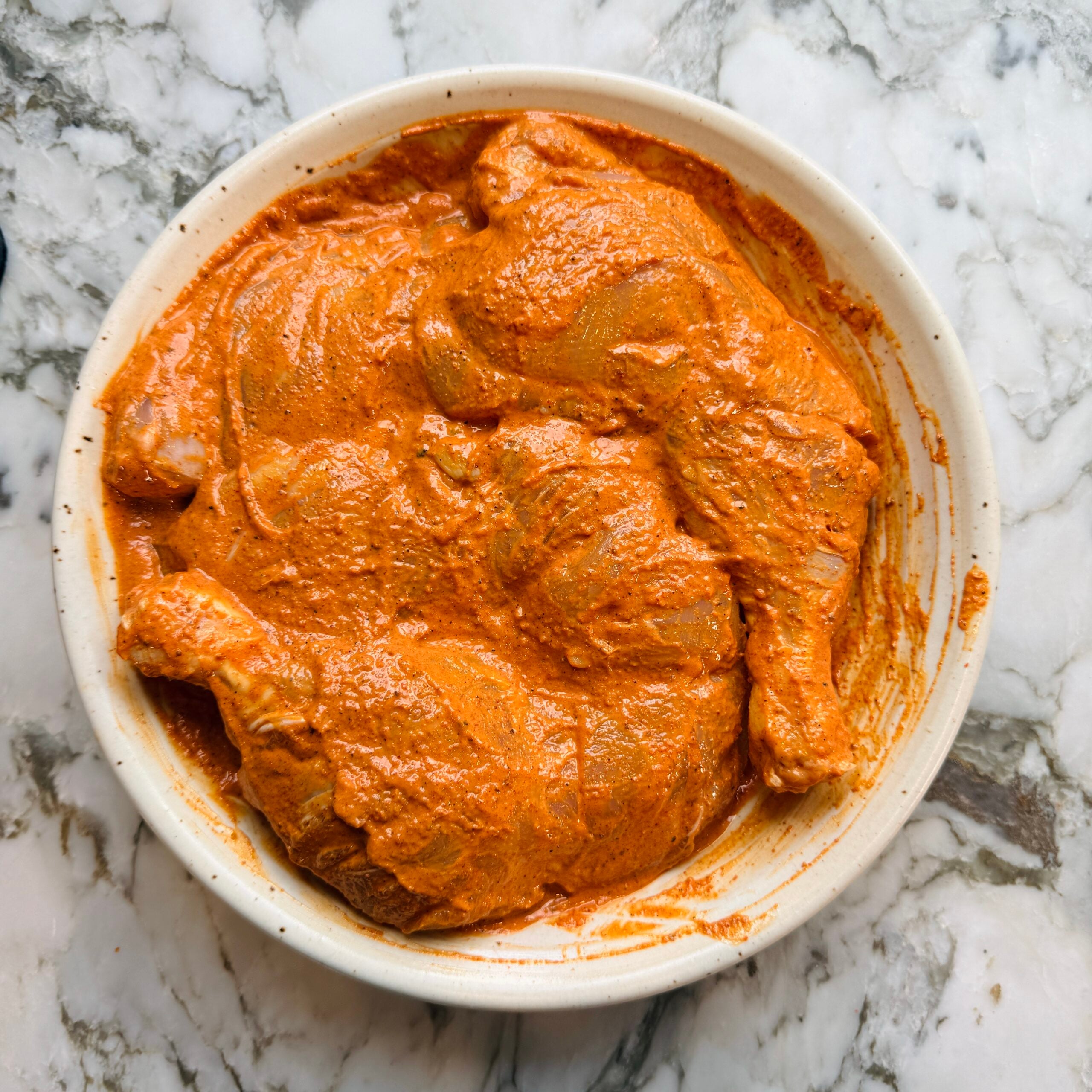 A bowl filled with raw chicken pieces coated in a thick, orange-red marinade sits on a marble surface.