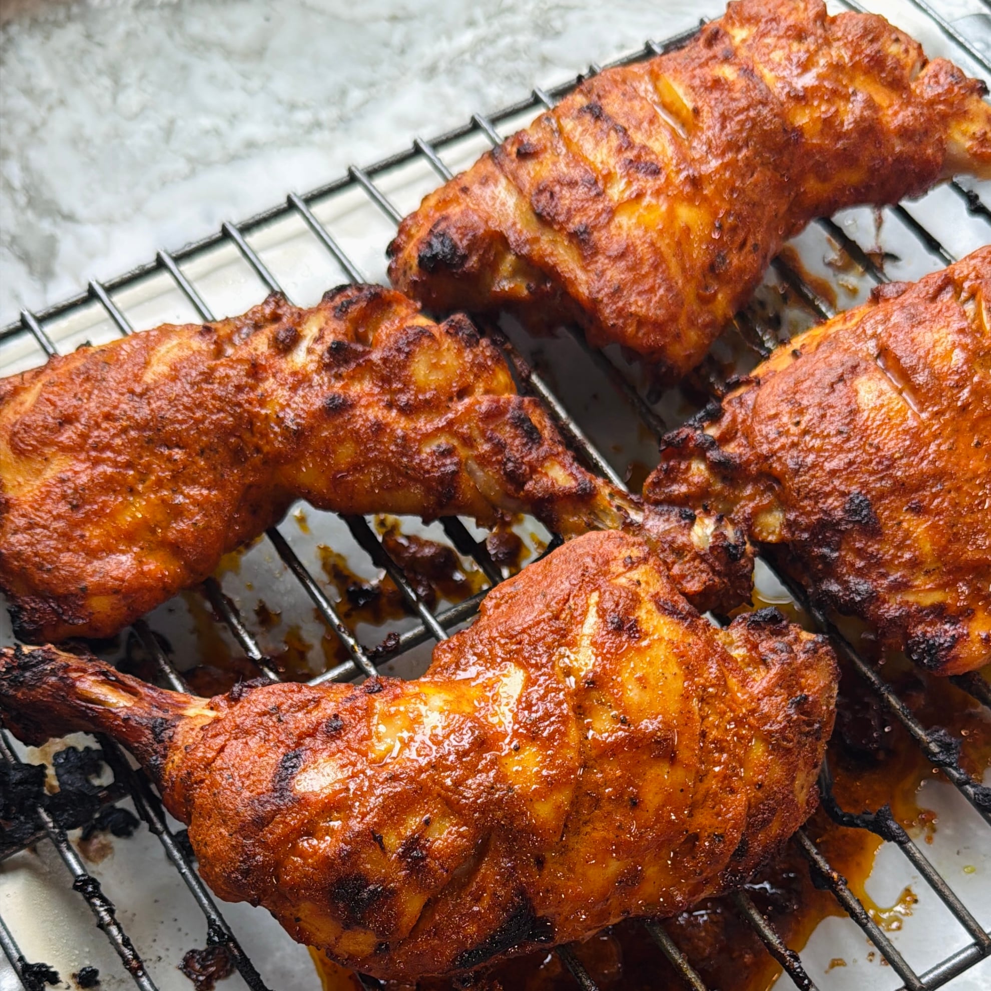 Four pieces of grilled, seasoned chicken with tandoori marinade are placed on a wire rack above a metal tray.