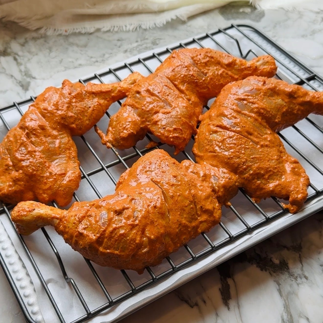Five marinated chicken legs coated in maronade on a wire rack over a baking tray on a marble countertop.