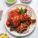 Three pieces of grilled tandoori chicken served on a plate with sliced red onions, fresh herbs, and lemon wedges on the side.
