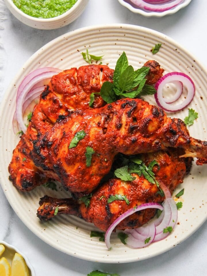 Three pieces of grilled tandoori chicken served on a plate with sliced red onions, fresh herbs, and lemon wedges on the side.